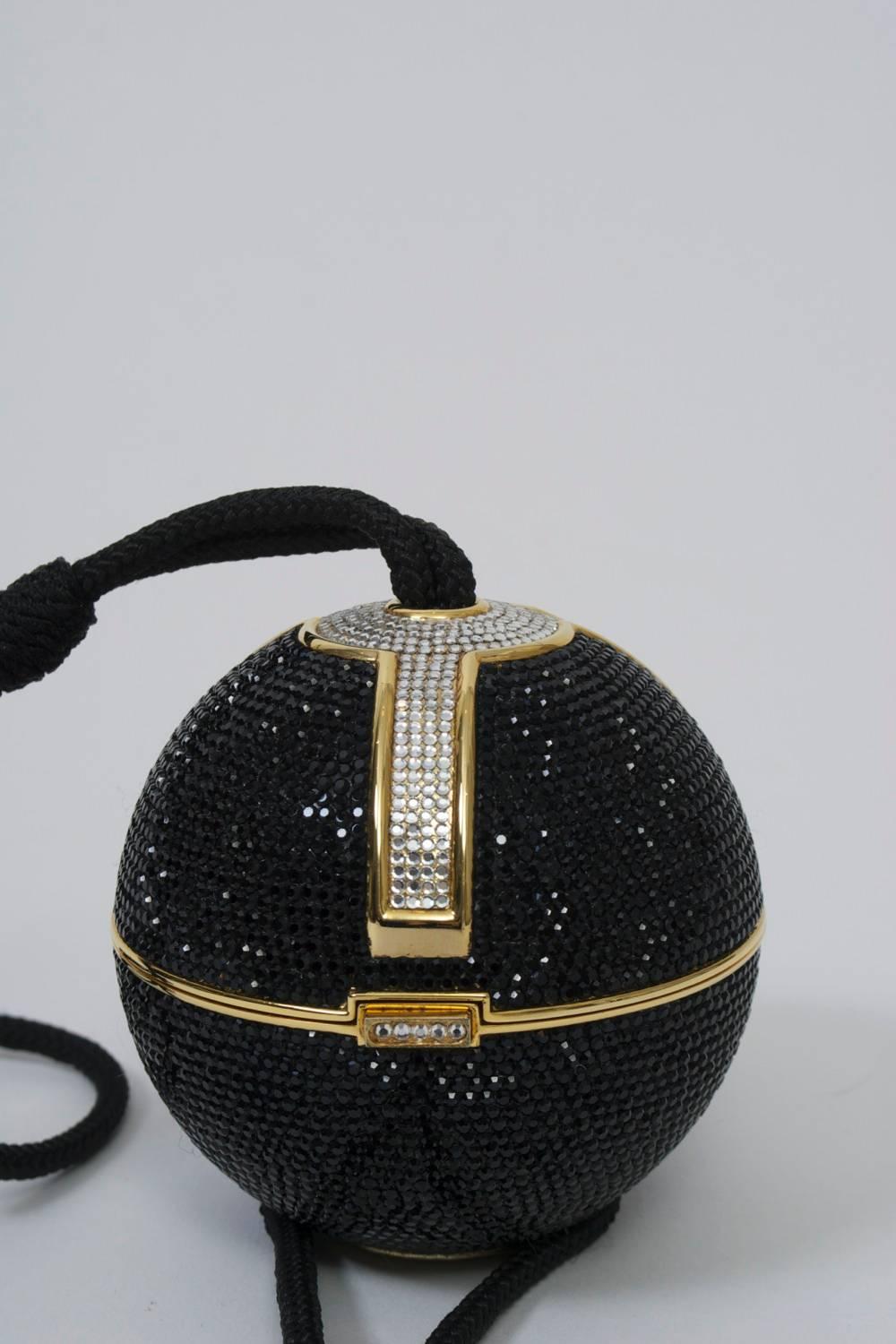 Judith Leiber spherical minaudiere covered in black pavé rhinestones punctuted with clear stones at top continuing down the front contained in a gold metal frame. Sphere opens in center, revealing a gold leather interior with change purse. Long