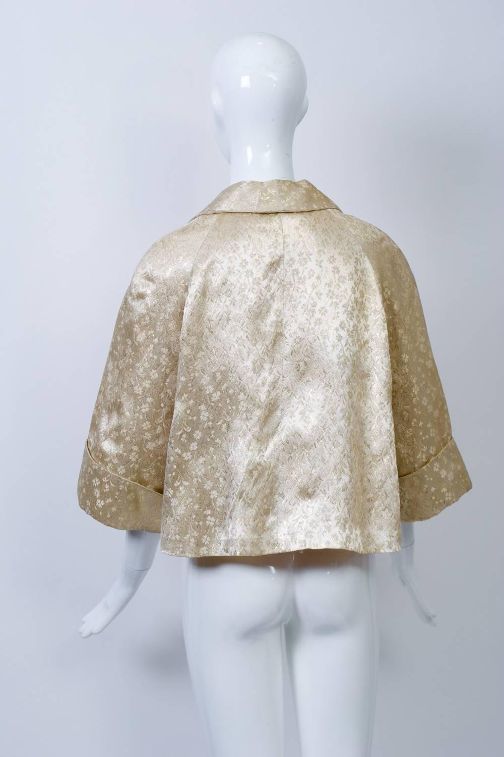 Vintage short swing jacket in ivory and gold brocade features wide, graduated lapels and wide three-quarter cuffed sleeves. Lined in beige taffeta. Retailed by B. Altman & Co. One size.