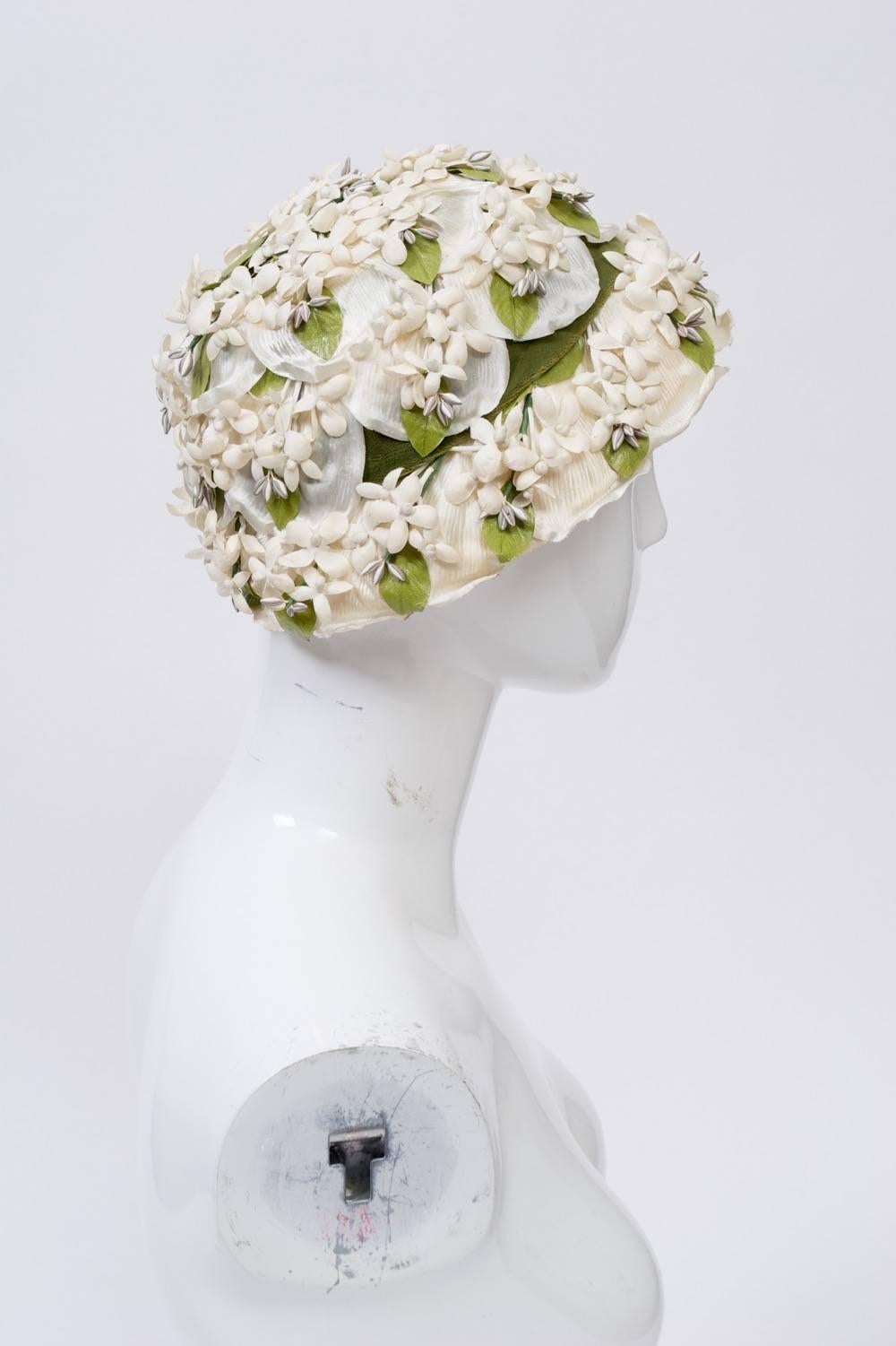 Small-brimmed 1950s hat decorated with clusters of white flowers and green leaves and finished with a green velvet band. Stiff lace structure underneath.
Retailed by Lord & Taylor. Size S-M.