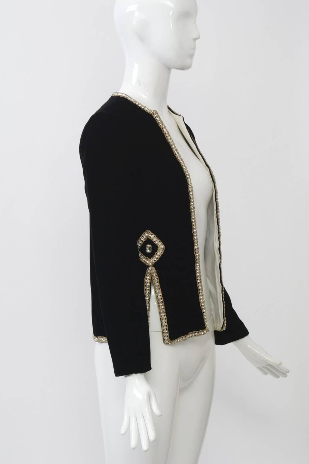 1960s Malcolm Starr black velvet features gold braid and rhinestone trim edging around the open front, round collar, and hem, as well as the deep slits towards each side, where they are topped by a diamond-patterned embellishment centering a large