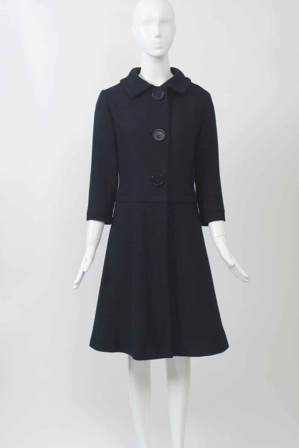 1960s lightweight coat by Jacques Heim in navy nubby wool featuring a fitted bodice and A-line skirt with a welted seam below the waist, three-quarter sleeves, and a free panel over the skirt in back, Spread collar and large buttons down the front.