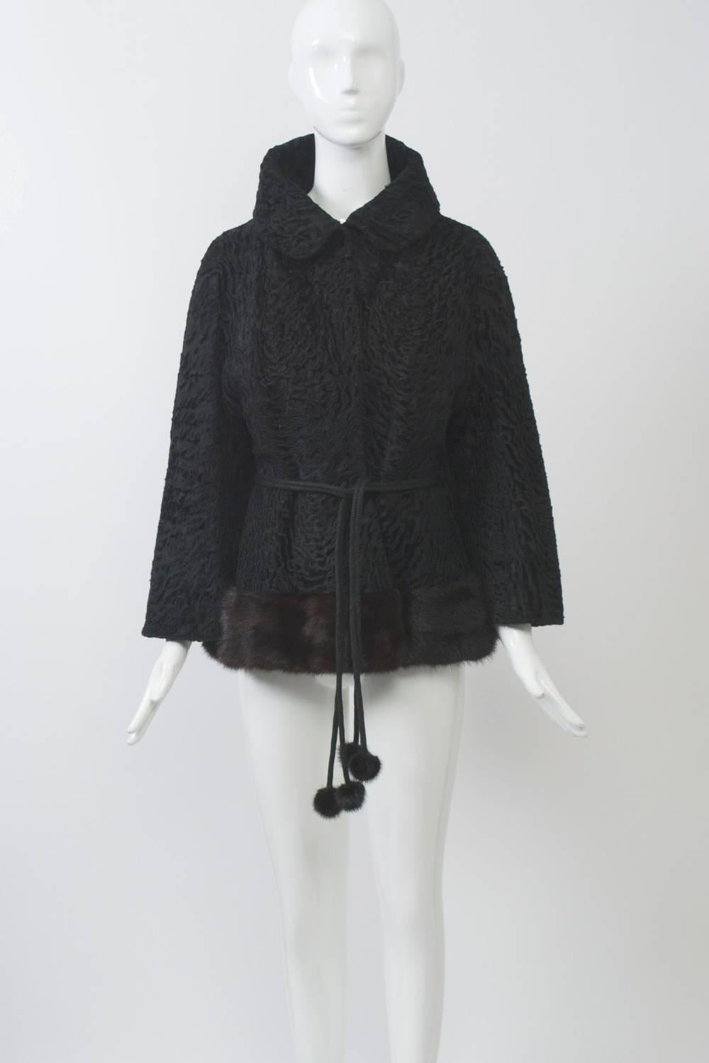 Black broadtail jacket features stand-up collar and deep ranch mink border along hem. A cord belt ending in mink pompoms goes through side slits and cinches the waist in front, leaving the back free. Very nicely and unusually styled. 
