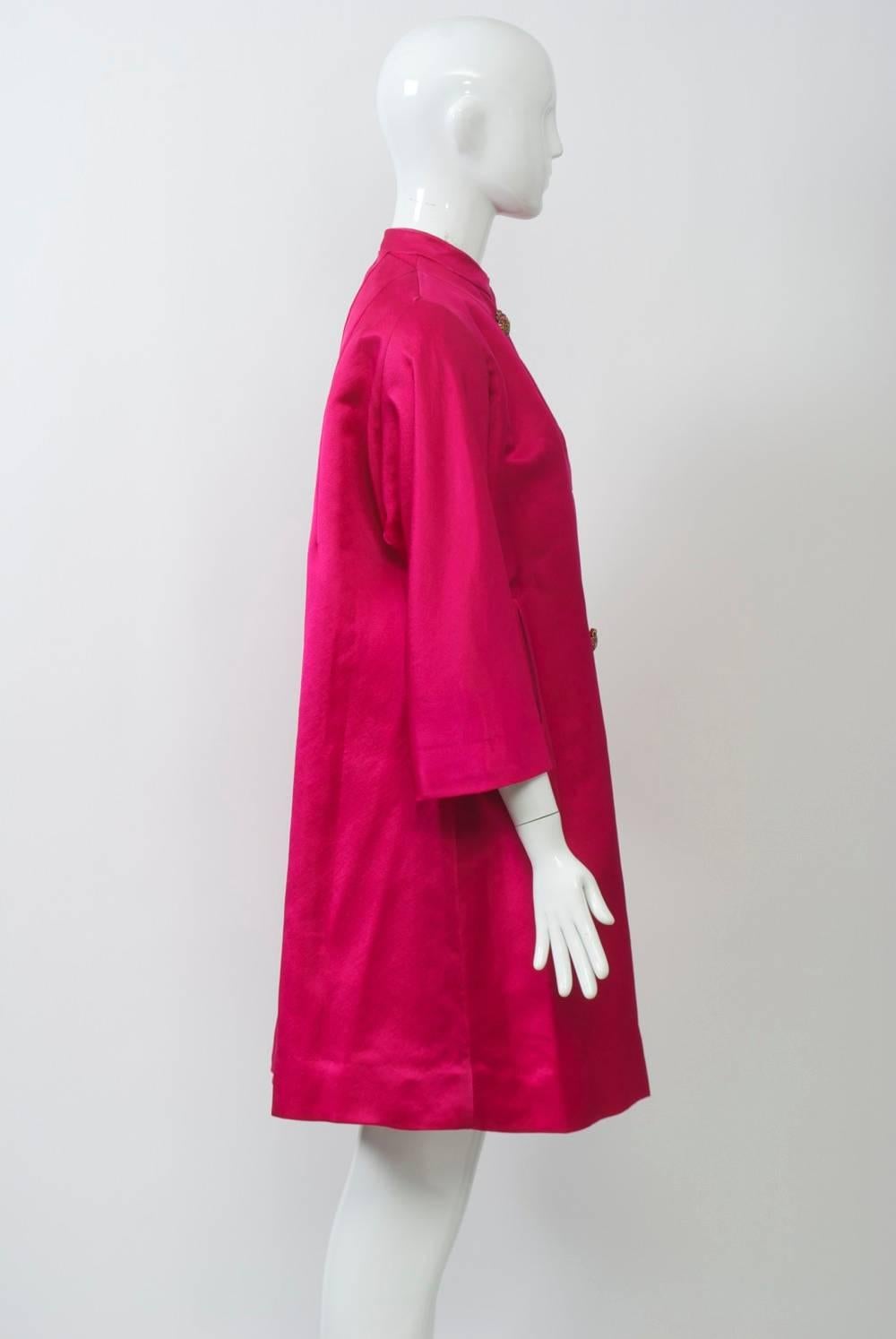 1960s silk evening coat in bright pink silk features an A-line profile with three-quarter sleeves and mandarin collar. Double-breasted, bound buttonholes with stone buttons and in inverted detailing at bust. Approximate size 6-8.

