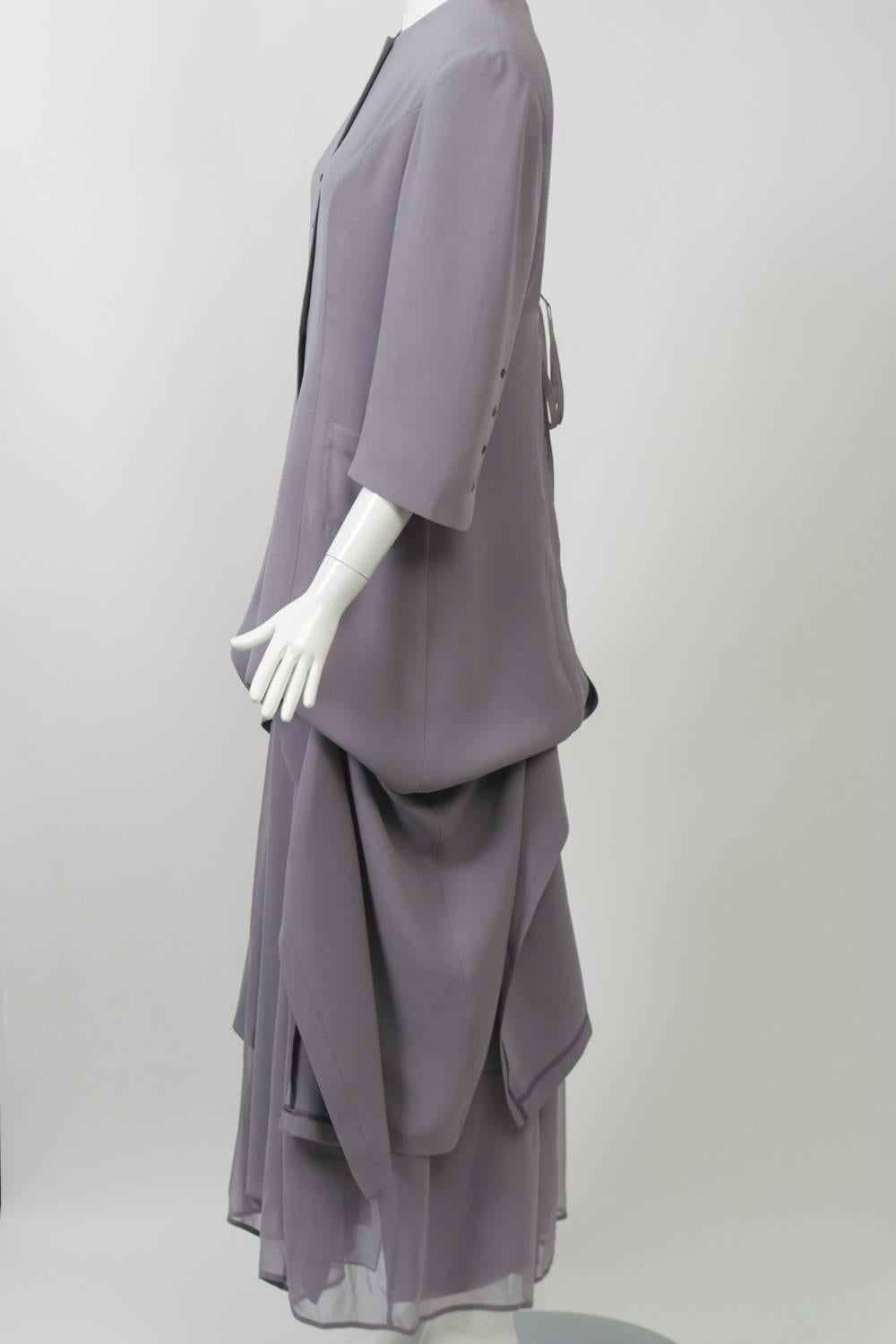 Morgane Le Fay long silk coat and dress in a subtle shade of lilac-tinged gray with intriguing detailing. The coat features interior ties, two on each side, which draw up the long skirt like a shade, revealing a layer of chiffon underneath, which is