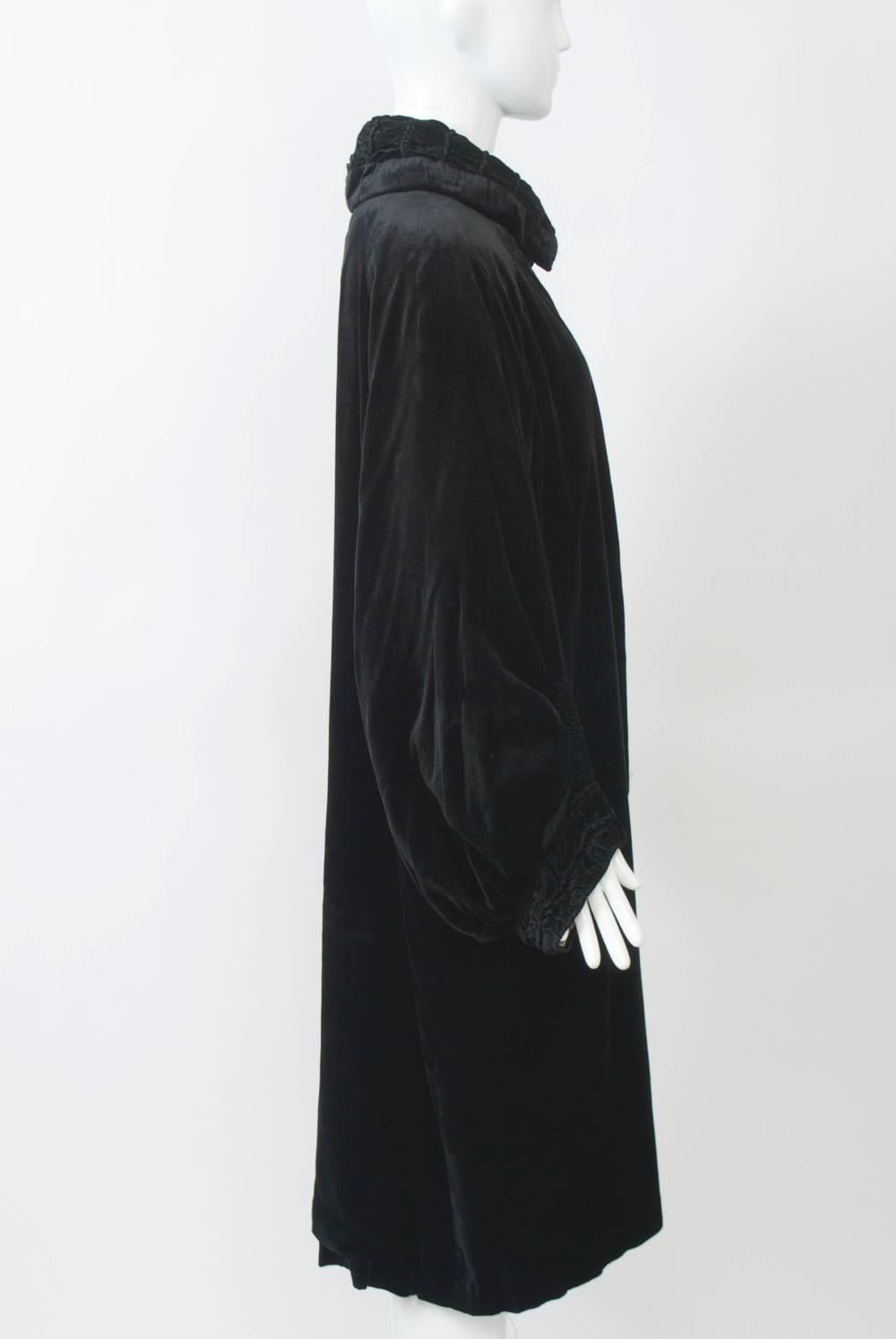 Black velvet opera coat featuring a ruched collar and wrists. Loose fit with curved front hem and balloon sleeves. Single rhinestone button closure. White satin lining.