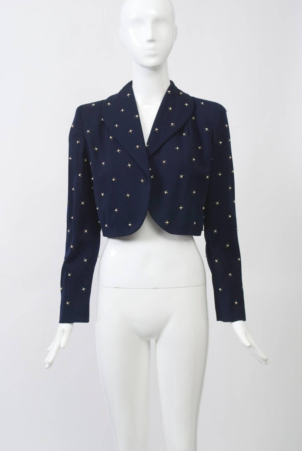 Cropped jacket in navy blue wool studded throughout with pearls and featuring a short shawl collar and curved front. Set-in narrow sleeves. Navy crepe lining. Approximate size 6.