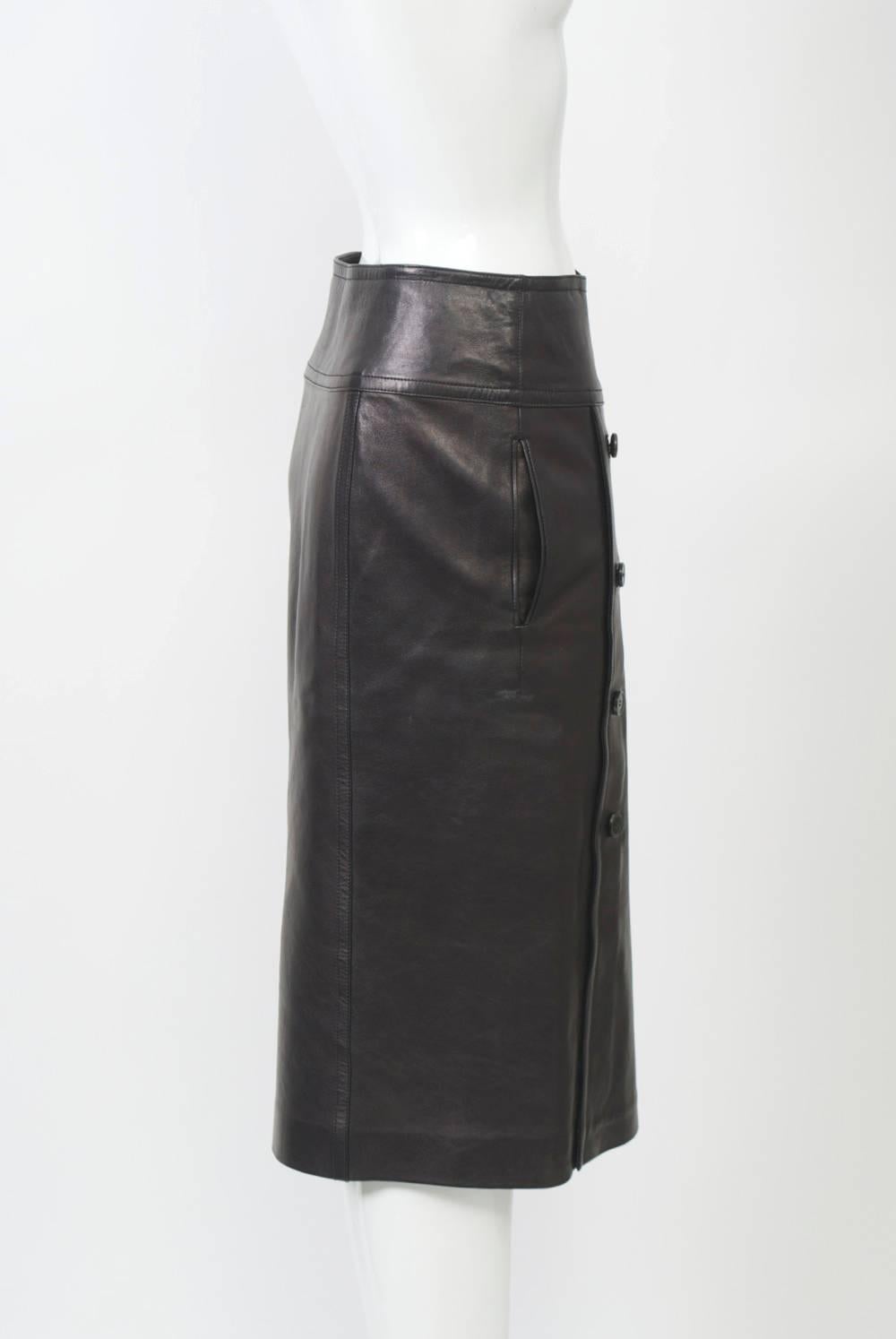 Black leather double-breasted slim skirt by Yves Saint Laurent with buckled hip band and side slit pockets. Below knee length, tapered profile.
