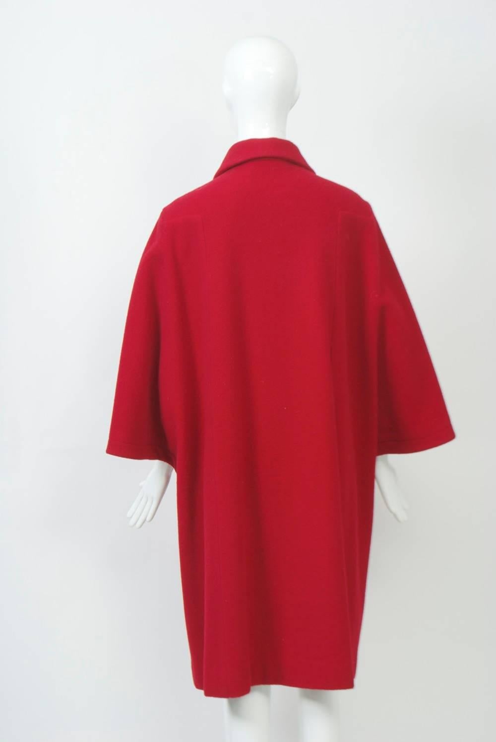 Striking red wool coat by Adolfo featuring cape sleeves, a small collar, and four black dome buttons down the front. Underneath the cape sleeves, the arms are free. Side pockets. Unlined. Interesting construction and detailing. Long black gloves