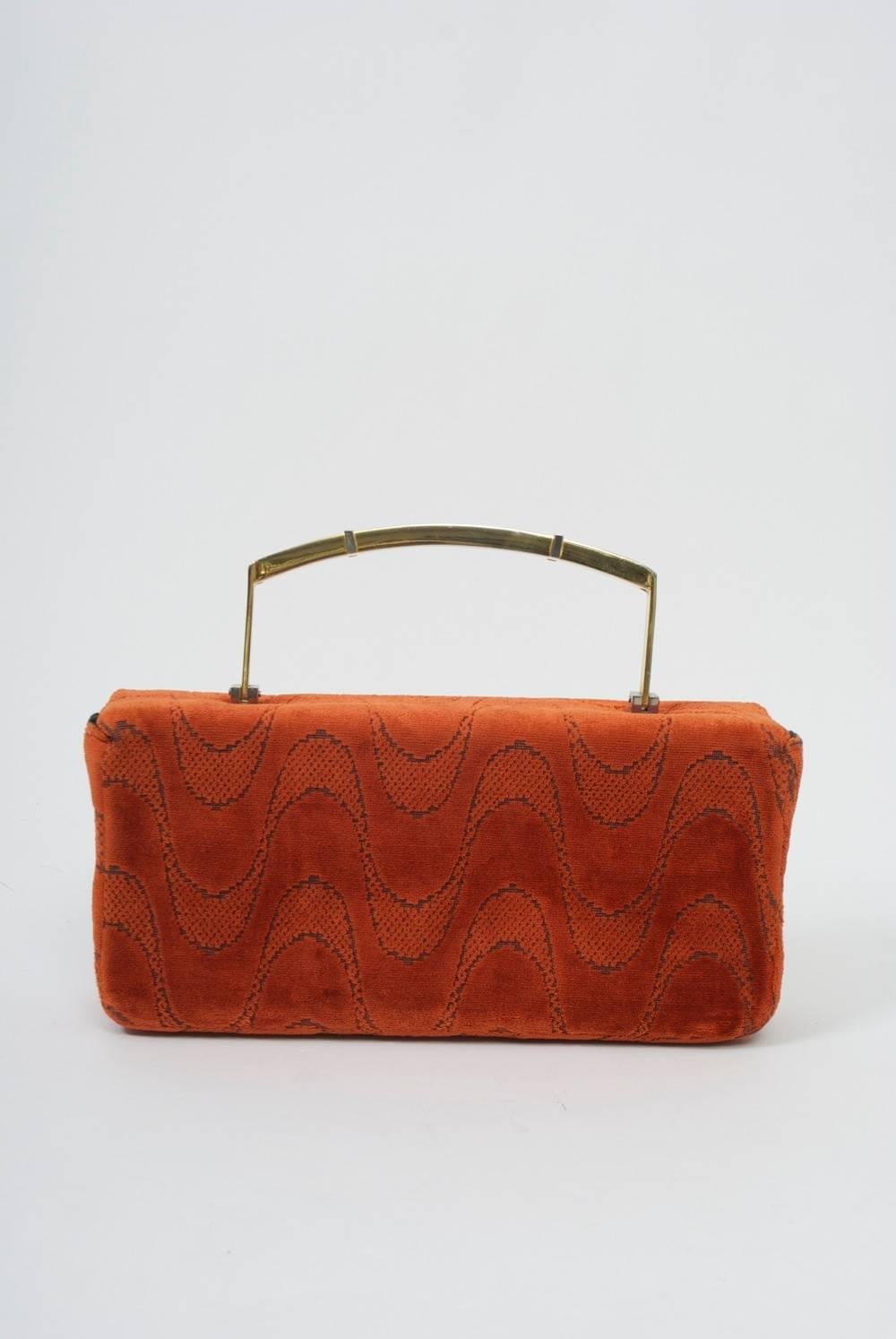 Unusual rectangular box bag in orange cut velvet with metal handle in gold accented in silver.The rectangular clasp pulls down to open the bag. Black faille interior with side compartments.