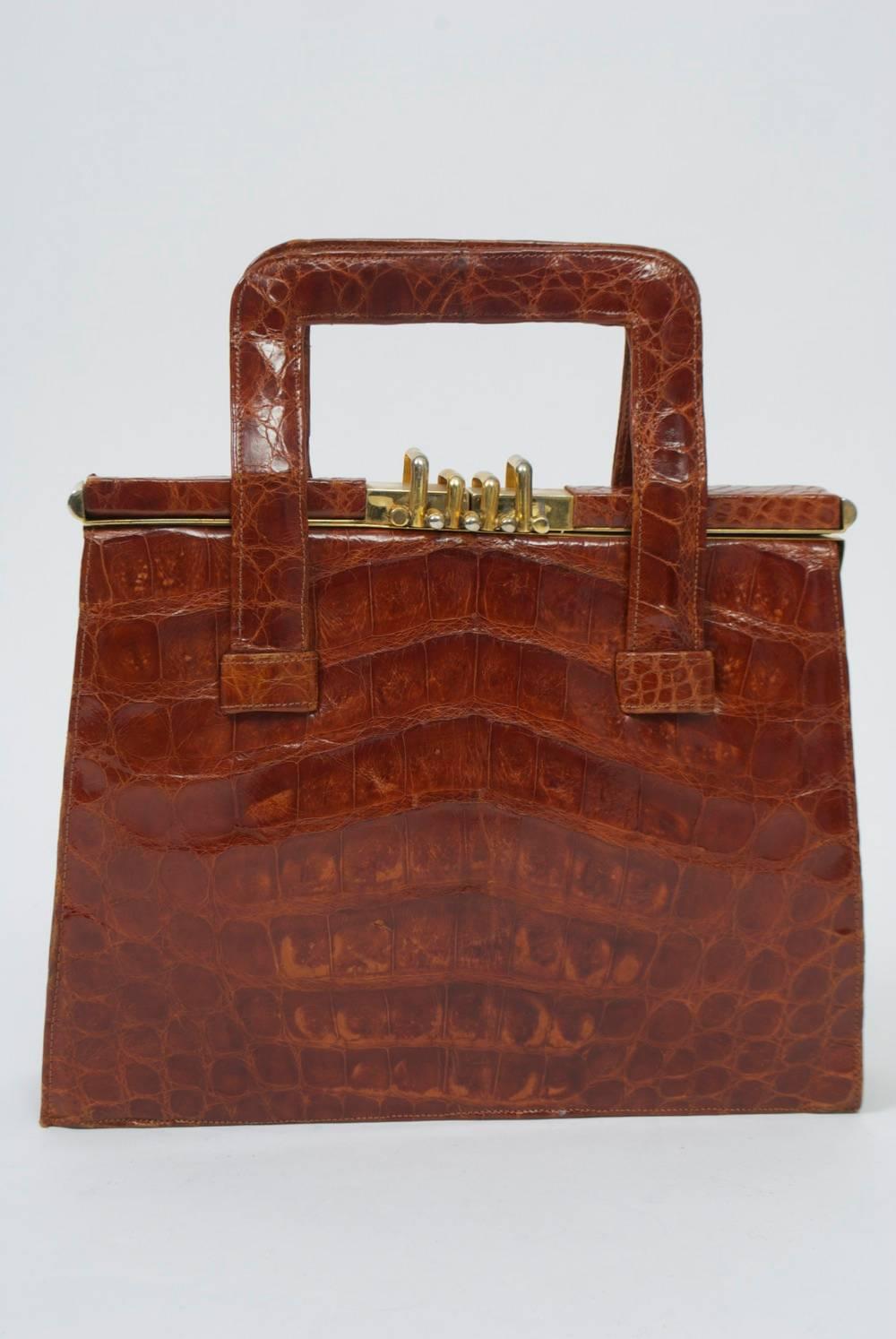 Alligator handbag, c.1950s, made in Argentina, in a rich shade of cognac. Structured, square shape with double handles and a gold metal frame partially covered in the alligator. At center, the unusual metal clasp pulls up to open the handbag. On the