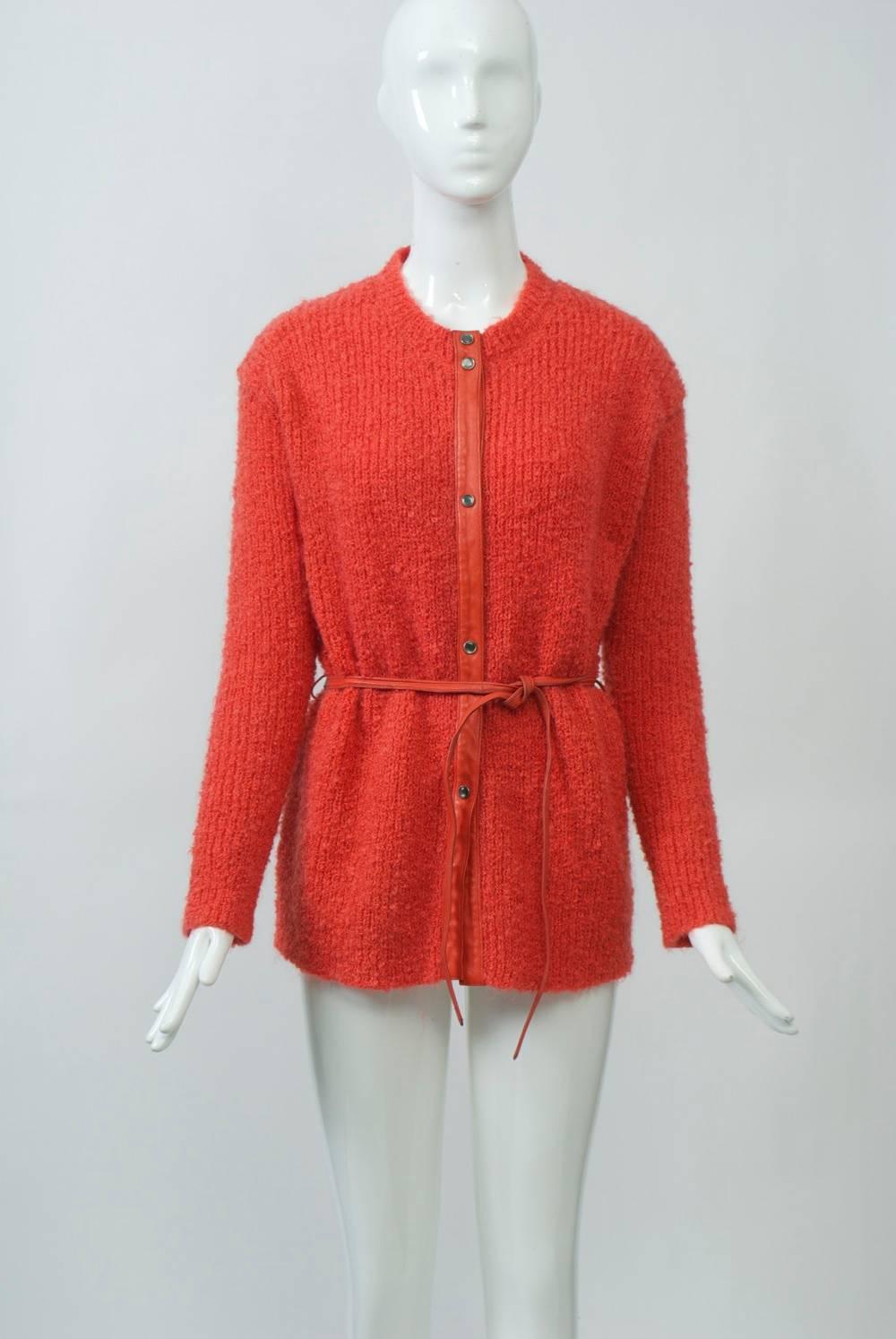 Cardigan in orange wool bouclé designed by Bonnie Cashin and featuring her characteristic leather trim down the front and in the narrow leather tie belt and gray pearl snap buttons. Orange leather also appears inside at the shoulders and central