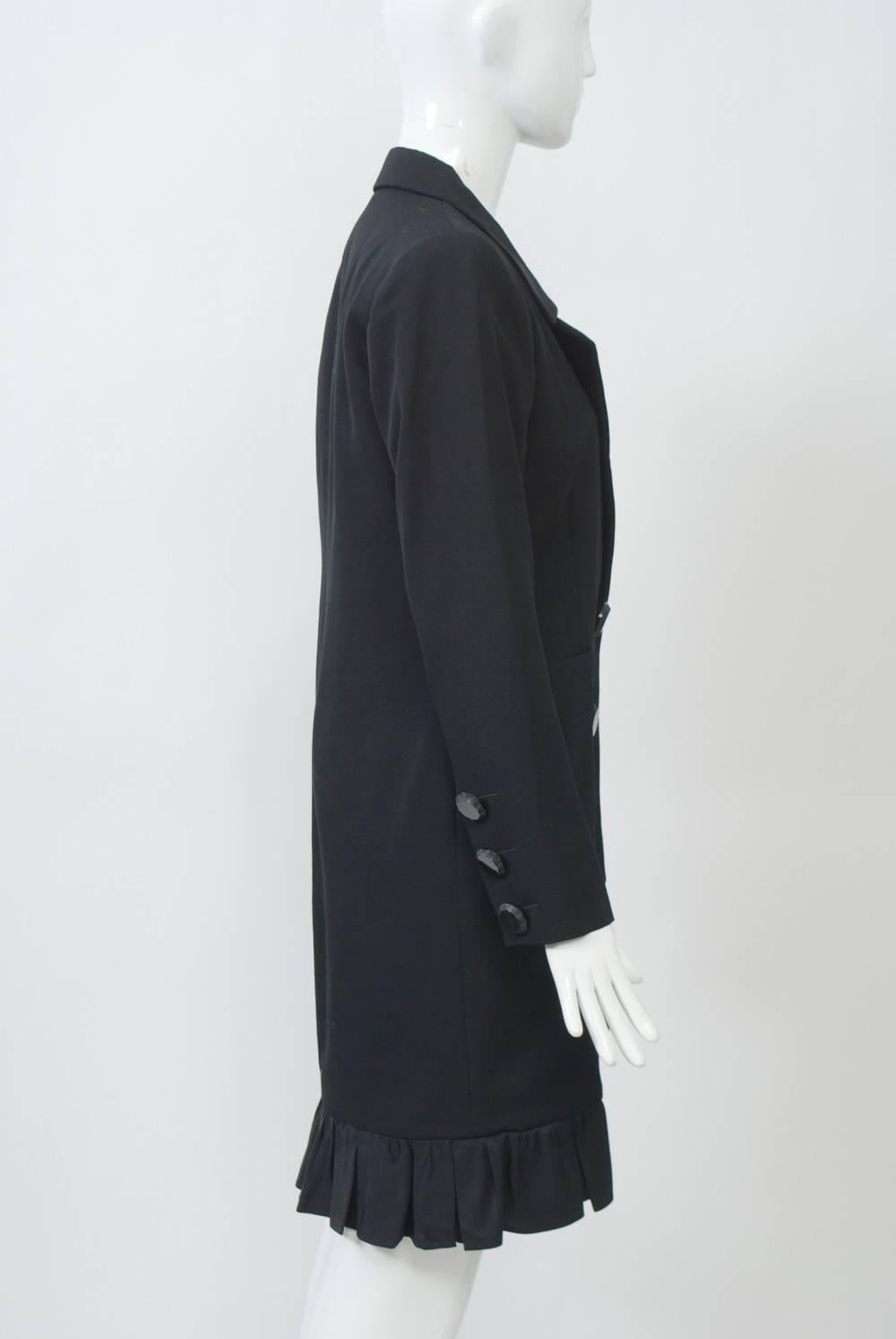 Black tuxedo-style coat dress with satin lapels and large jet buttons down front and at wrist. Satin ruffle at knee-length hem curves up at front. Shaped body with horizontal pockets.  Square shoulders with pads. Lined.