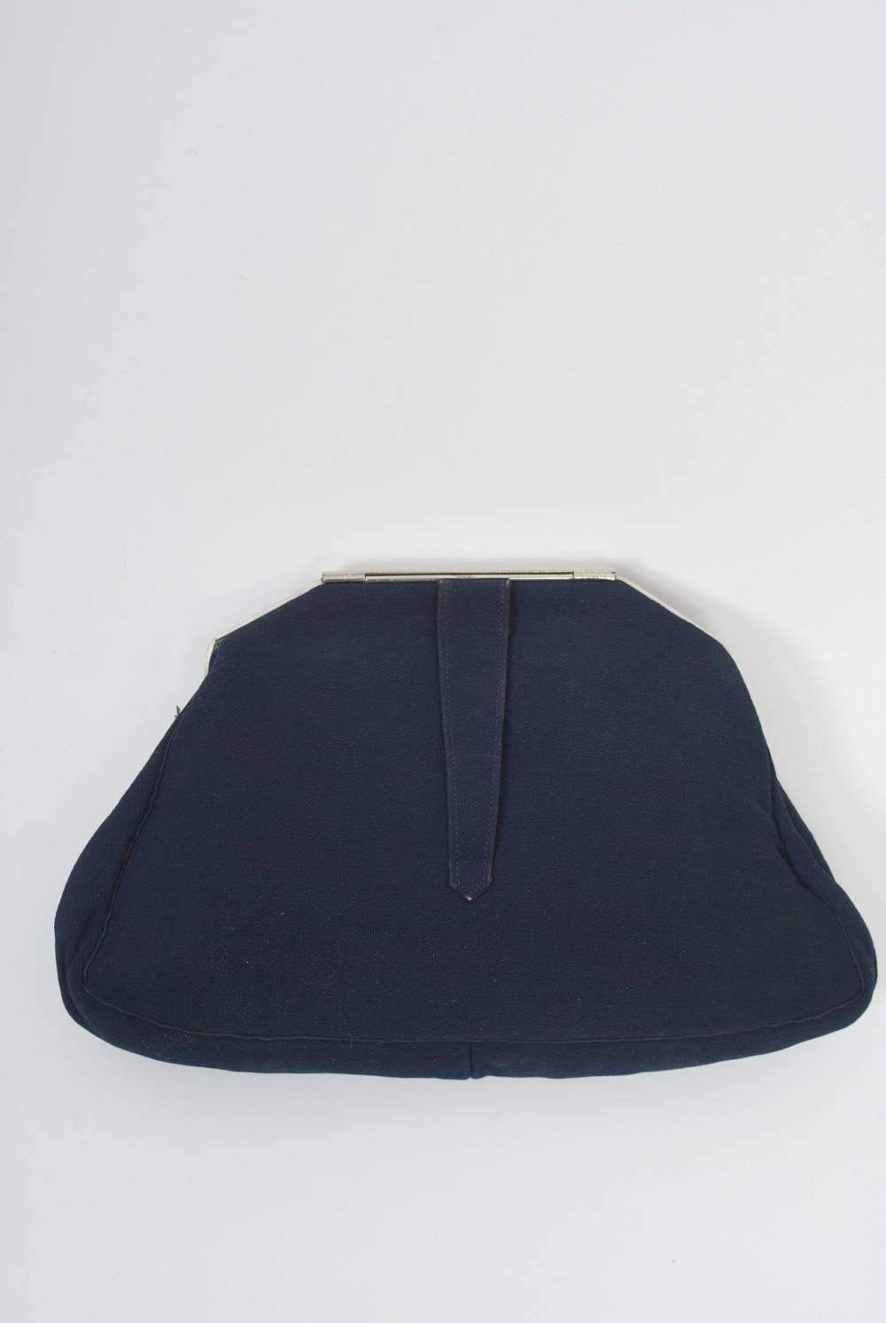 Evening clutch in navy silk featuring a silver metal frame and clasp, the latter quarter-moon shaped cut metal, the rounded edge framed in small rhinestones. A loop handhold is on the reverse of the clutch. The matching interior has an open side
