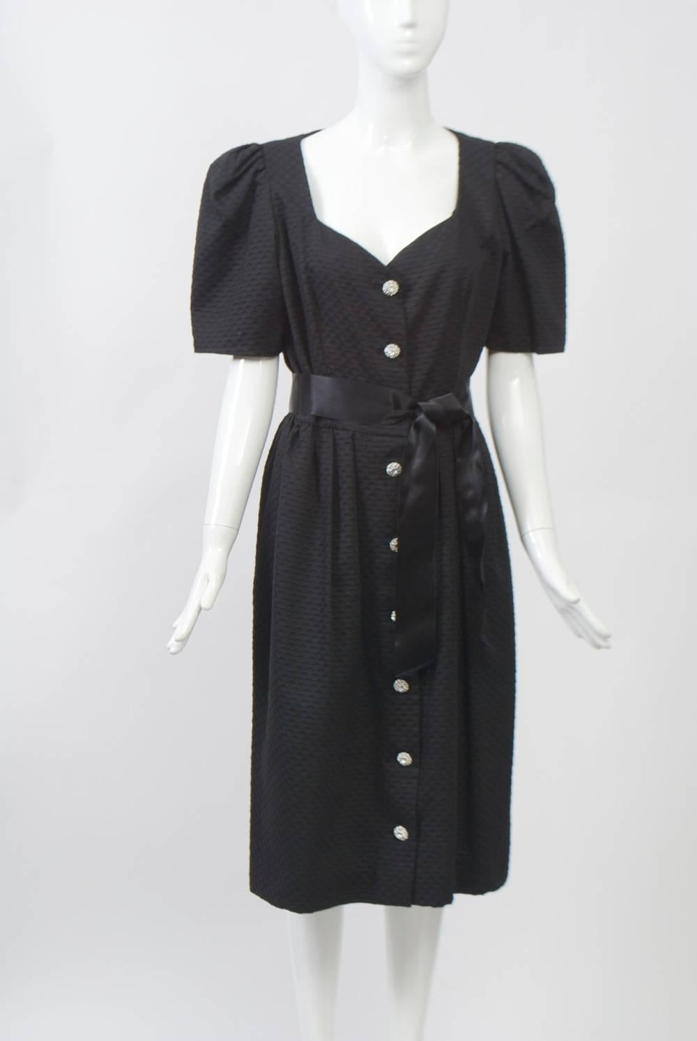 Shirt-style dress by Oscar de la Renta in black textured cotton dressed up with rhinestone buttons and a satin sash, as well as a sweetheart neckline. Additional features are puffed, short sleeves and a gathered skirt that ends below the knees.