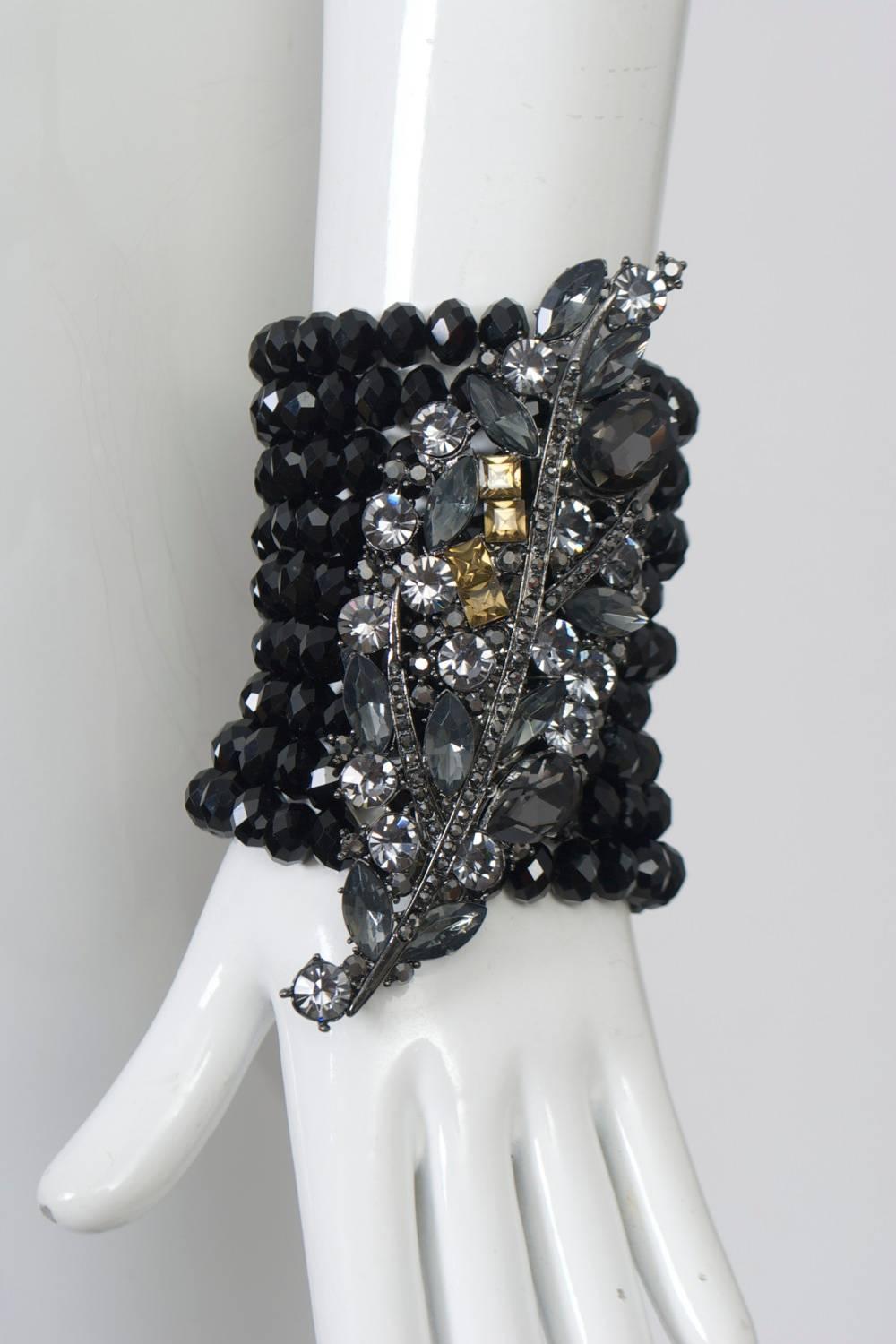 A dramatic bracelet composed of nine rows of faceted black beads on stretch cords featuring a large centerpiece of leaf form set with light and dark gray crystals of various shapes and sizes accented by four golden-toned stones. Toby Fischer, under