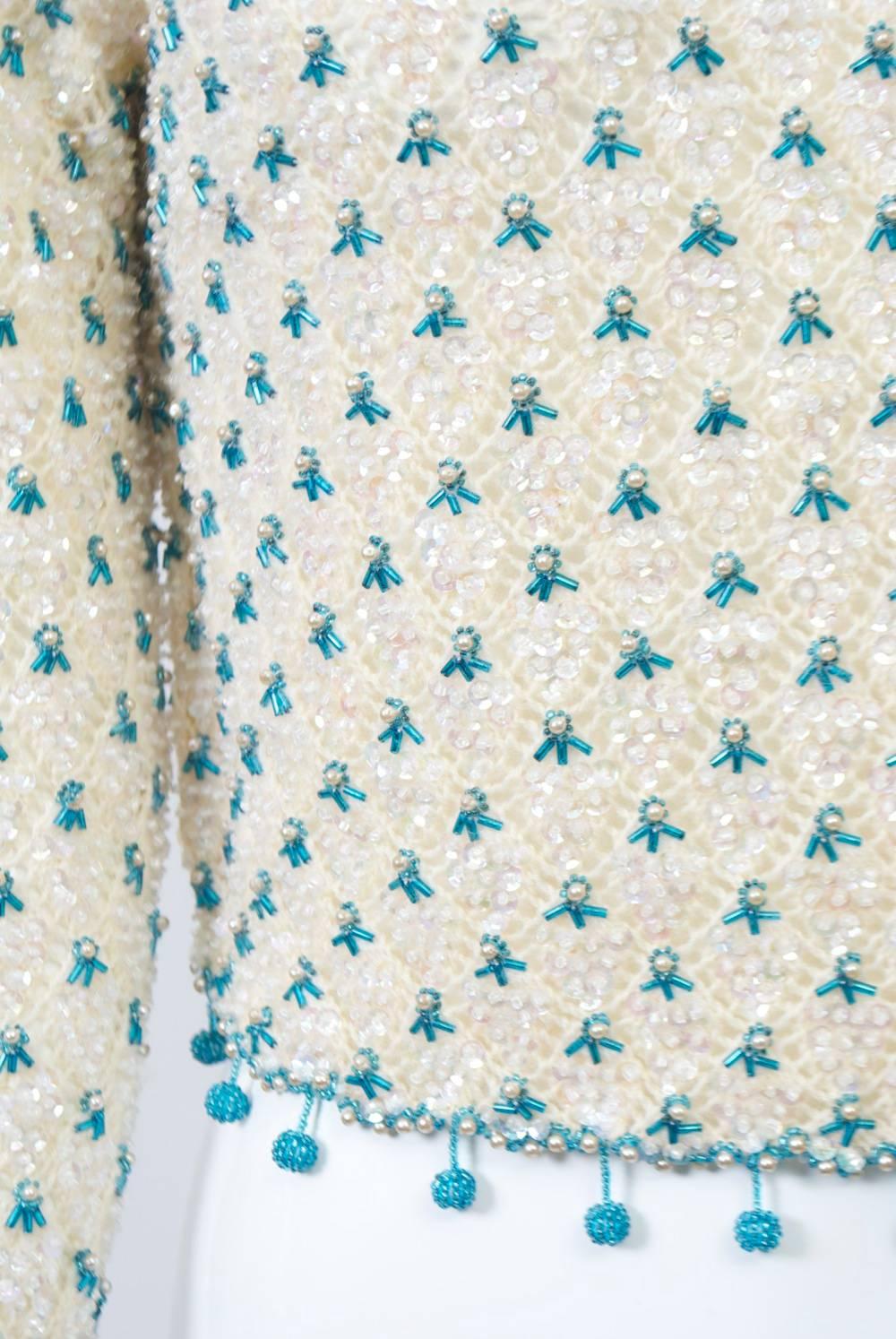 1950s-'60s cropped ivory sweater covered in ABA sequins and studded throughout in a turquoise bead and pearl motif; turquoise beaded edging is further adorned with turquoise beaded balls around the waist. The cropped knit features an open design, a
