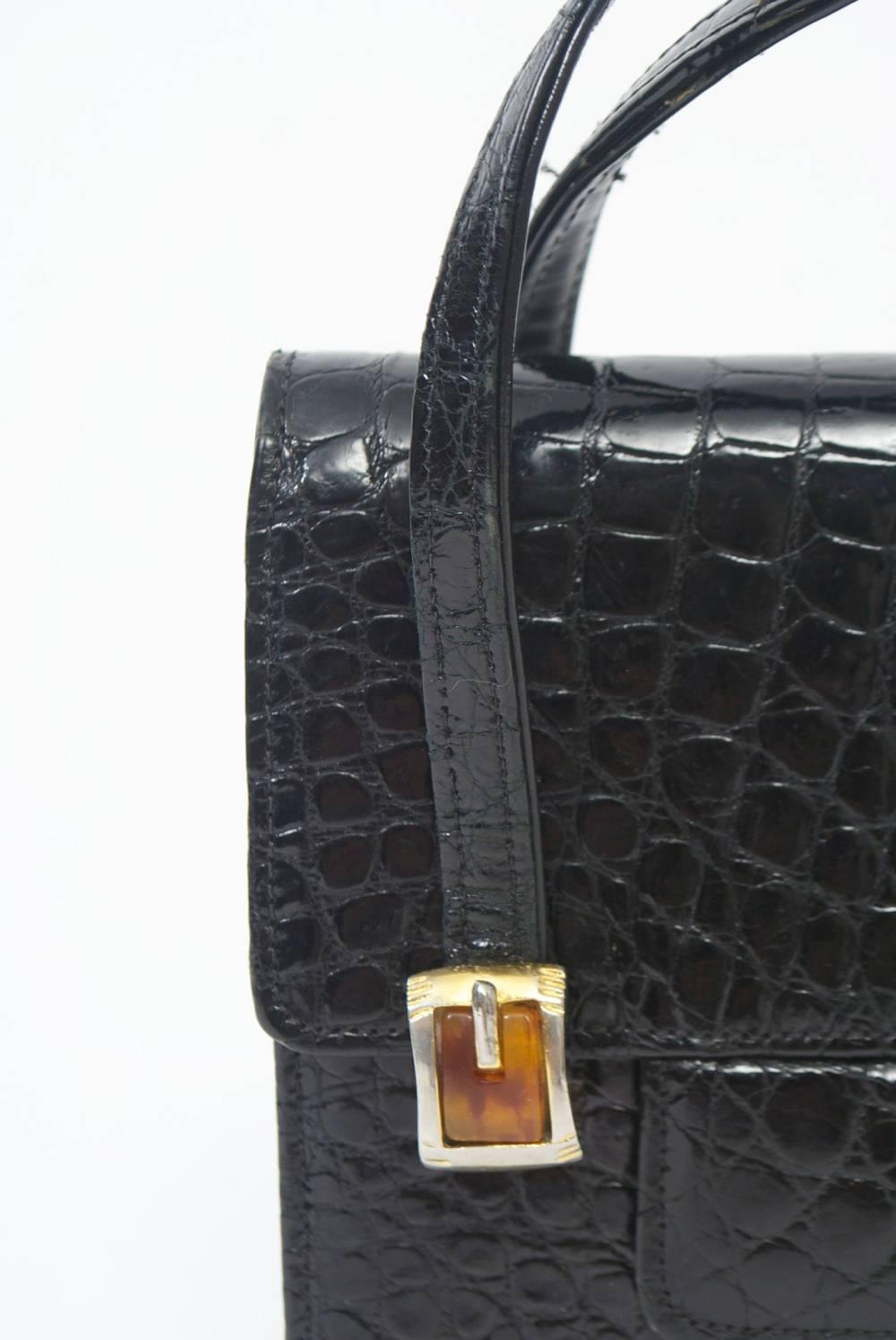 1960s black alligator handbag featuring a foldover flap and double handles secured in front with small enameled buckles. Accordion interior with zippered compartment on one side. 