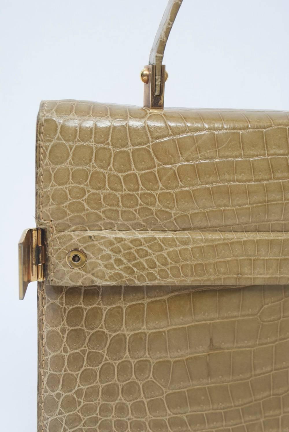 Great size and neutral color define this alligator handbag featuring gold metal clasps on either side of the envelope flap. The single handle is fastened with similar metal braces. On the beige leather interior, there are multiple pockets, including