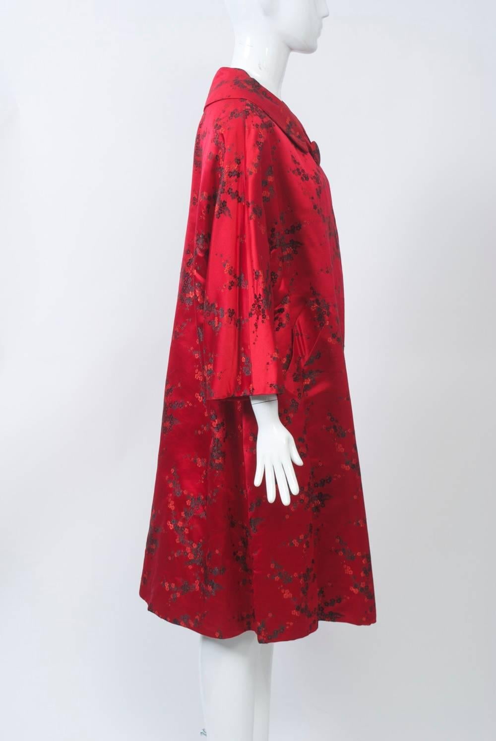 Red satin brocade evening coat with dainty black floral design featuring a spread collar and one large self button at the neck. Dolman, three-quarter sleeves. Easy fit, slash pockets. Red satin lining. Looks unworn.