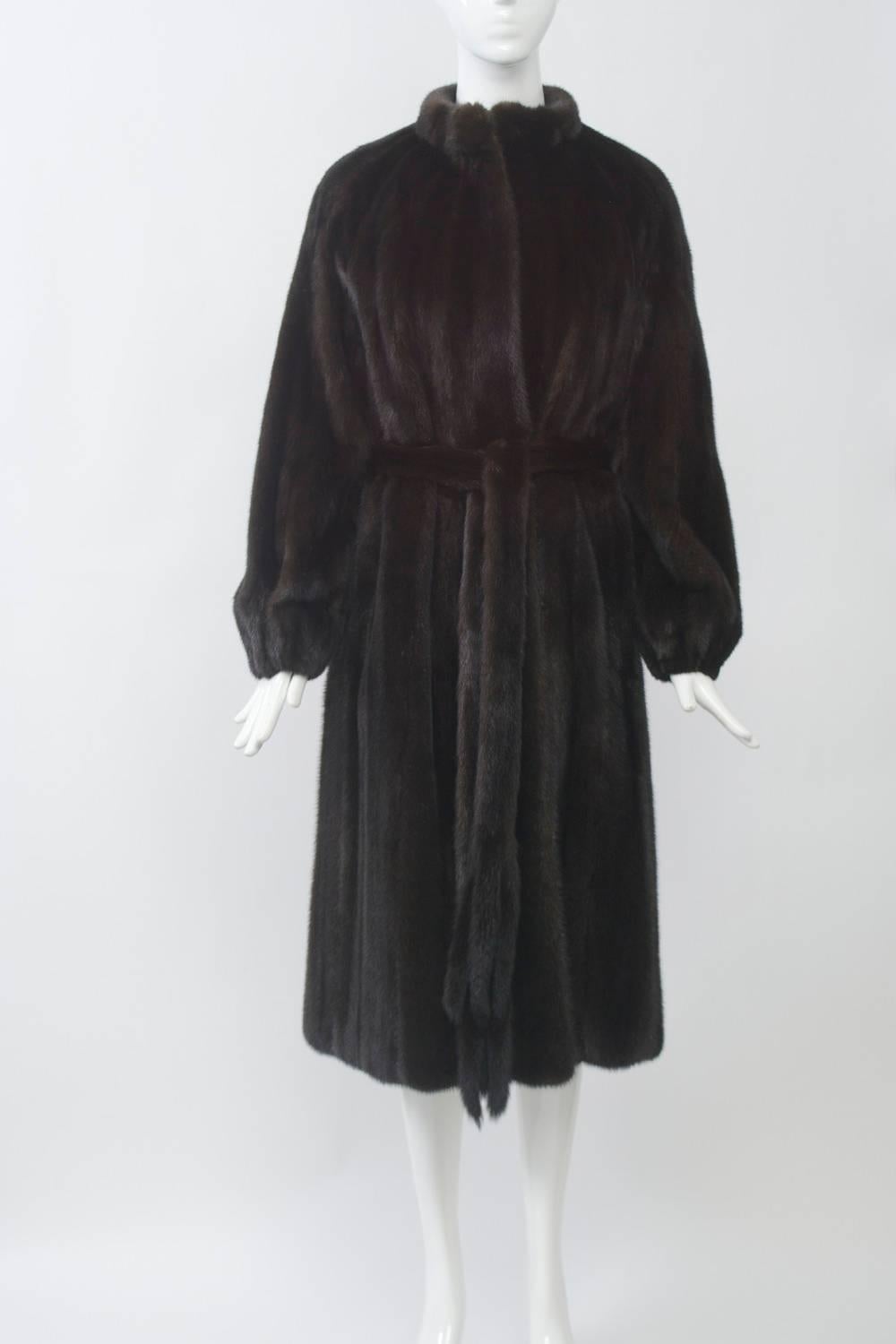 A classic, timeless mink coat of mahogany color and simple styling that features a slim body with modified A-line skirt and a funnel collar. Slit pockets. The coat is held close to the body with a long self mink tie terminating in tails. No
