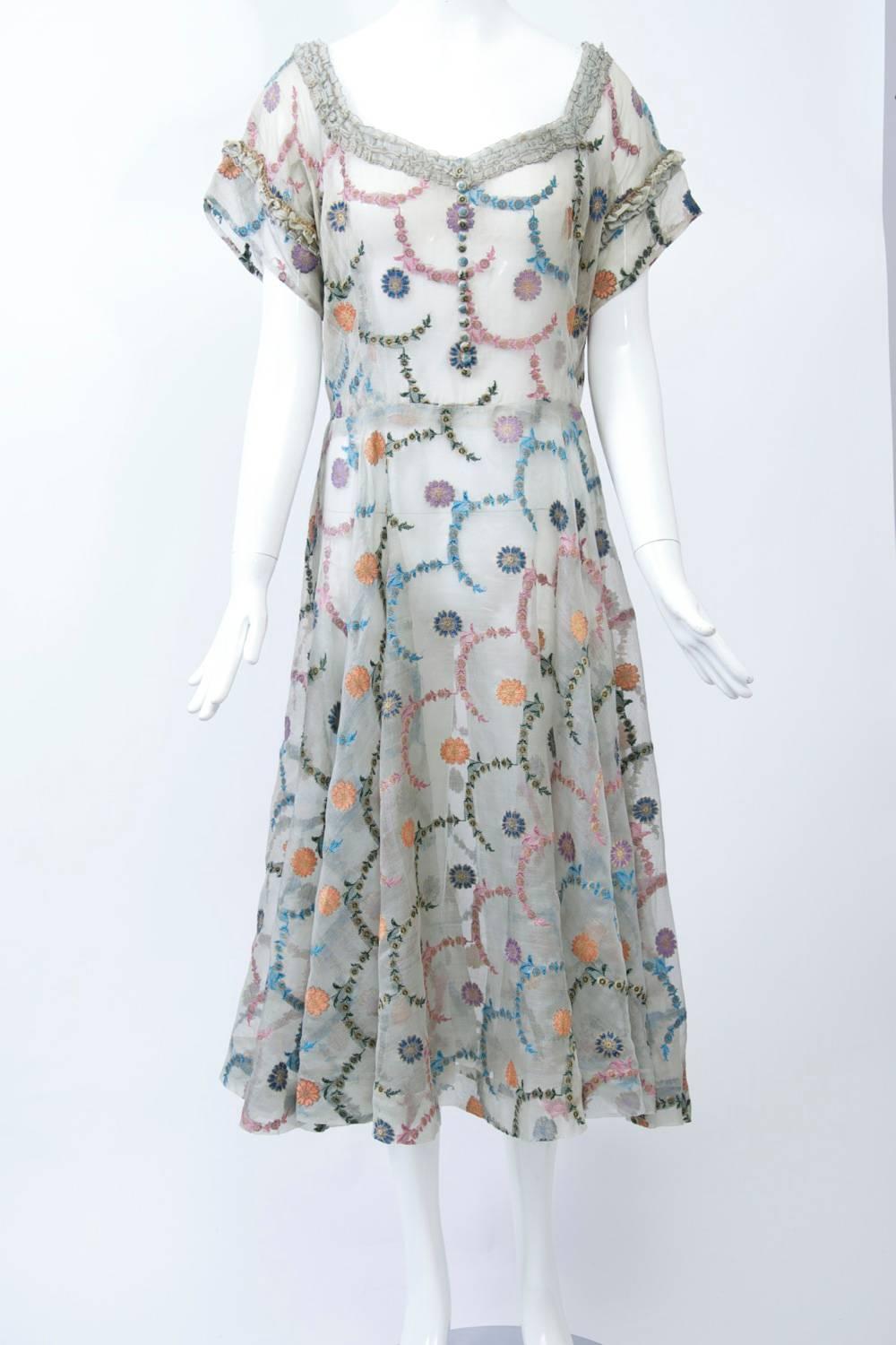 1950s silhouette in pale gray organza embroidered throughout with small flowers and half circle chains of smaller flowers and leaves in a pastel palette. The fitted bodice features short sleeves and ruching around the sweetheart neckline; the