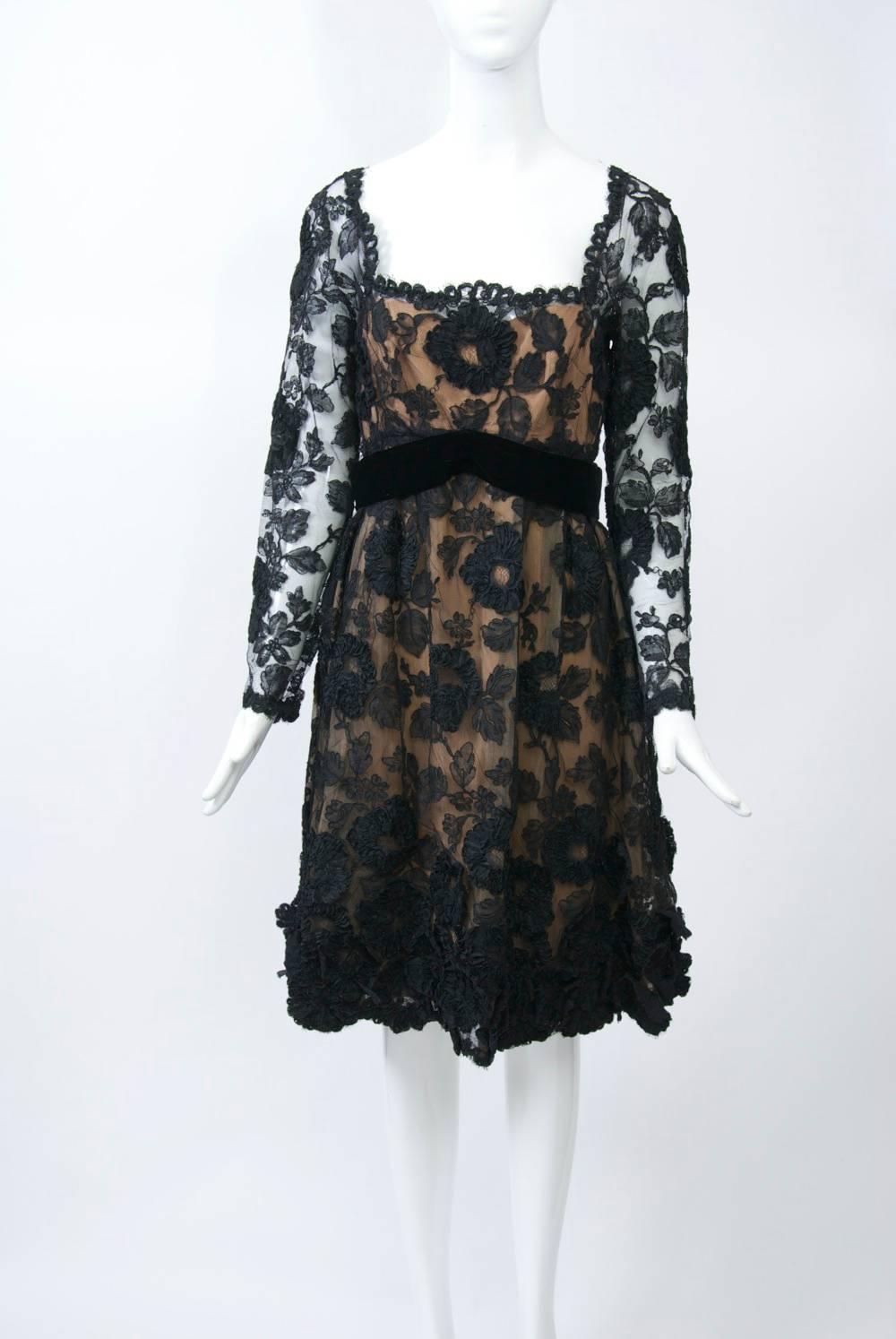 Vintage cocktail dress of black lace featuring a wide, square neckline, empire waist, and long sleeves.  Accents of black ribbon flower appliqués adorn the skirt, which has a scalloped hemline as does the neck and shoulders; a black velvet sash with