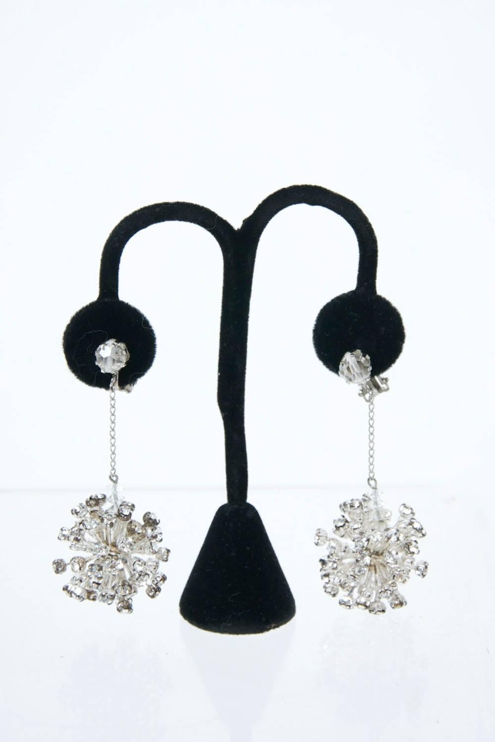 1960s atomic drop earrings, each sphere consisting of varied lengths of protrusions terminating in a rhinestone. The spheres are suspended by a thin silver chain from a single rhinestone earpiece. Clip ons. Unmarked.