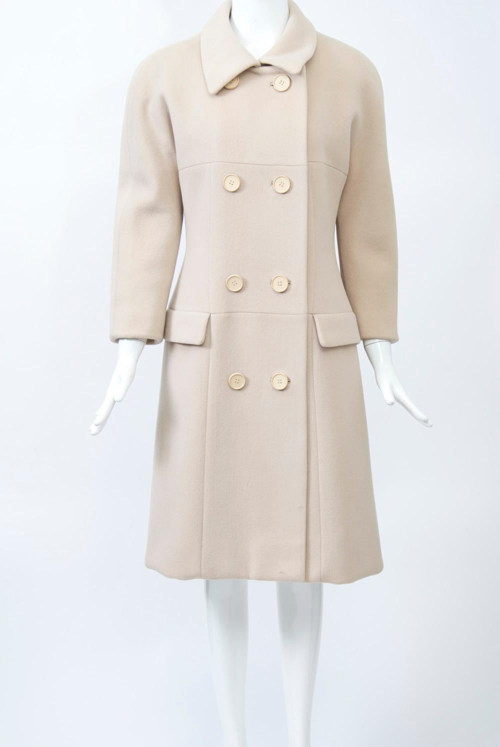 Iconic Norell 1960s styling defines this beige wool coat. Double breasted styling with eight buttons, a small pointed collar, and dolman sleeves that reach above the wrists. Special details include horizontal seams at the bust and the hip, where two