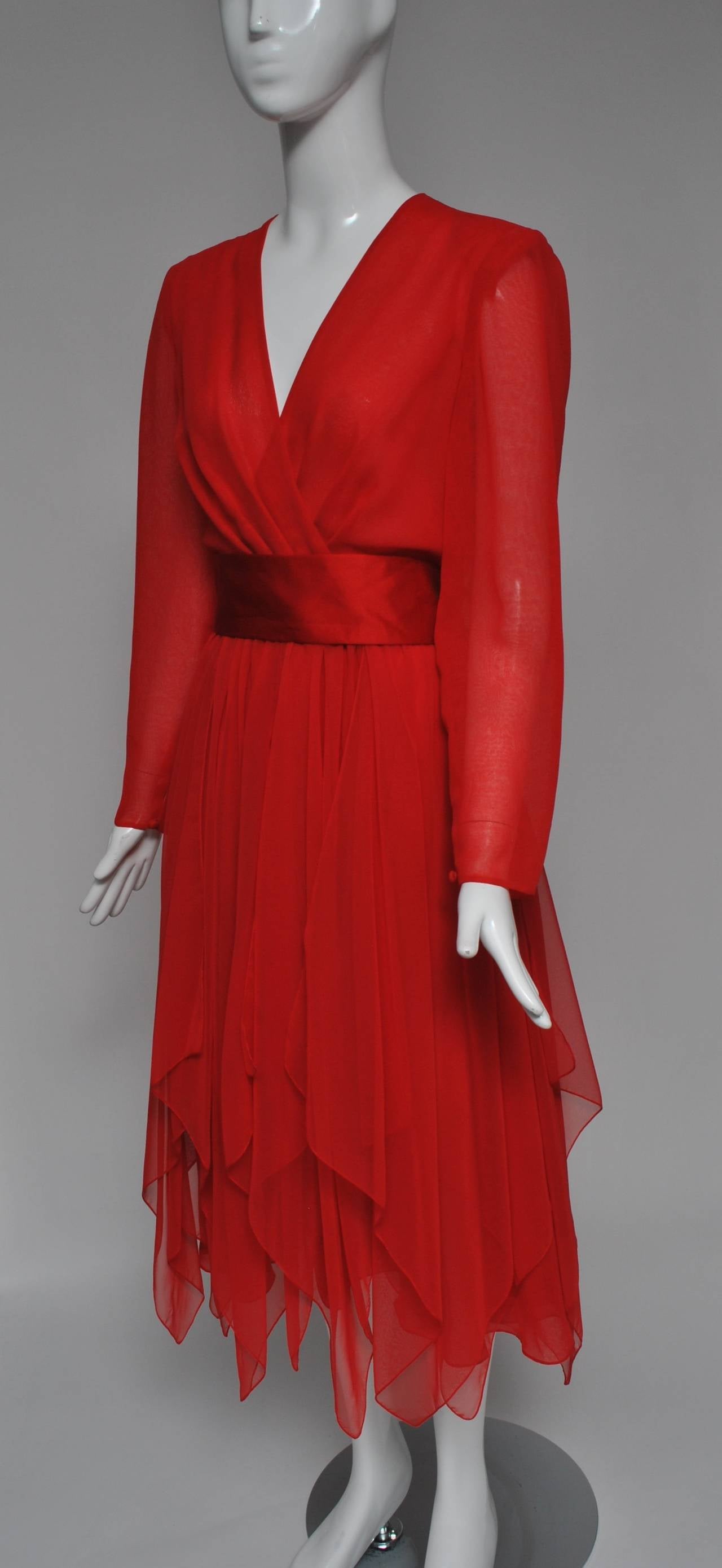 Late 1970s red cocktail dress in polyester chiffon by Estevez features a surplice top and handkerchief-paneled skirt. The long sheer sleeves button at the wrist. A wide red satin sash wraps around the waist and ties in back with long streamers.