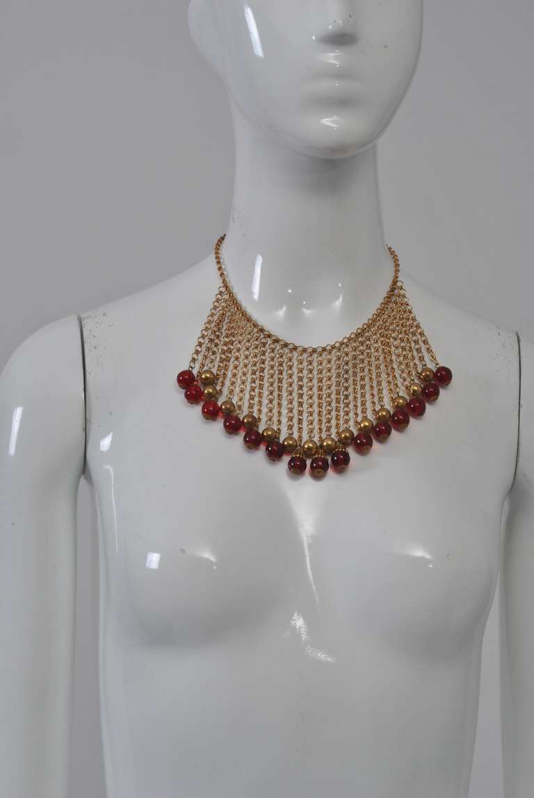 Bib necklace composed of small gold chains suspended from a neck chain, alternating between a shorter length terminating in a gold bead and a slightly longer length terminating in a red glass bead. A great look on the skin as well as on a high