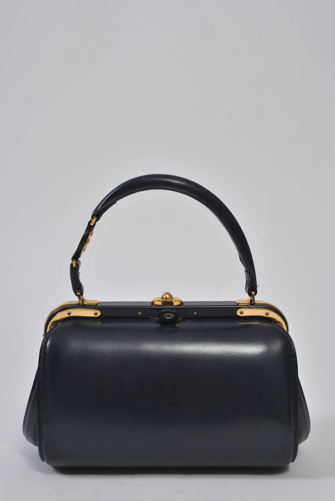 From high-end leather goods purveyor Lederer, this private-label 1960s handbag is crafted in France of dark navy leather and features distinctive gold metal hardware on the frame, handle, clasp, and feet. The rounded box shape is classic mid century