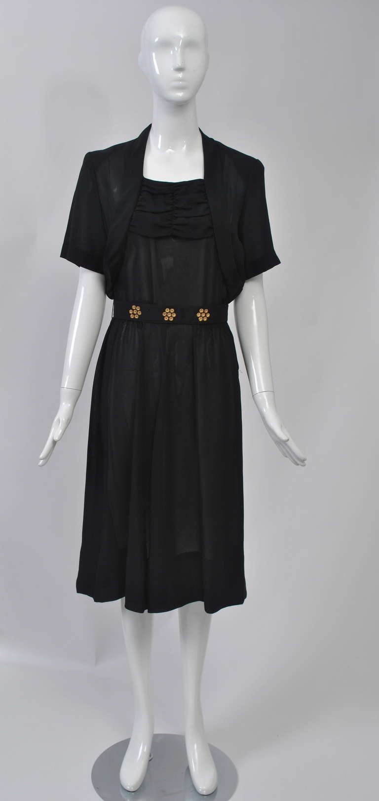 Black 1940s dress in a slightly sheer fabric, possibly rayon. The bodice, with short sleeves, has a bolero-like effect and shirring across the top. The gently shirred skirt falls softly. A belt that snaps in back has clusters of gold studs.