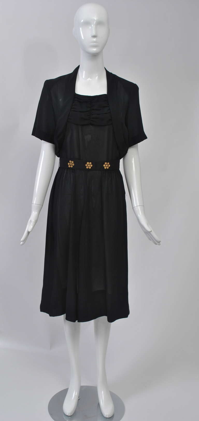 Robe noire des années 1940 en tissu légèrement transparent, probablement de la rayonne. Le corsage, à manches courtes, présente un effet de boléros et des fronces sur le dessus. La jupe légèrement froncée tombe doucement. Une ceinture qui se ferme