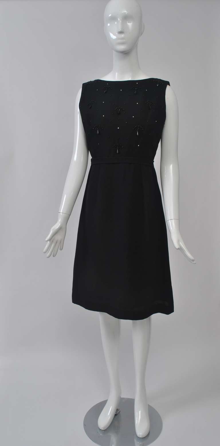LBD from the 1960s in crepe featuring a high-waisted bodice decorated with fan-shaped black beads and interspersed with rhinestones. A-line skirt with soft, wide pleats at back, waistband finished with bow. Straight neckline in front, scoop in back,