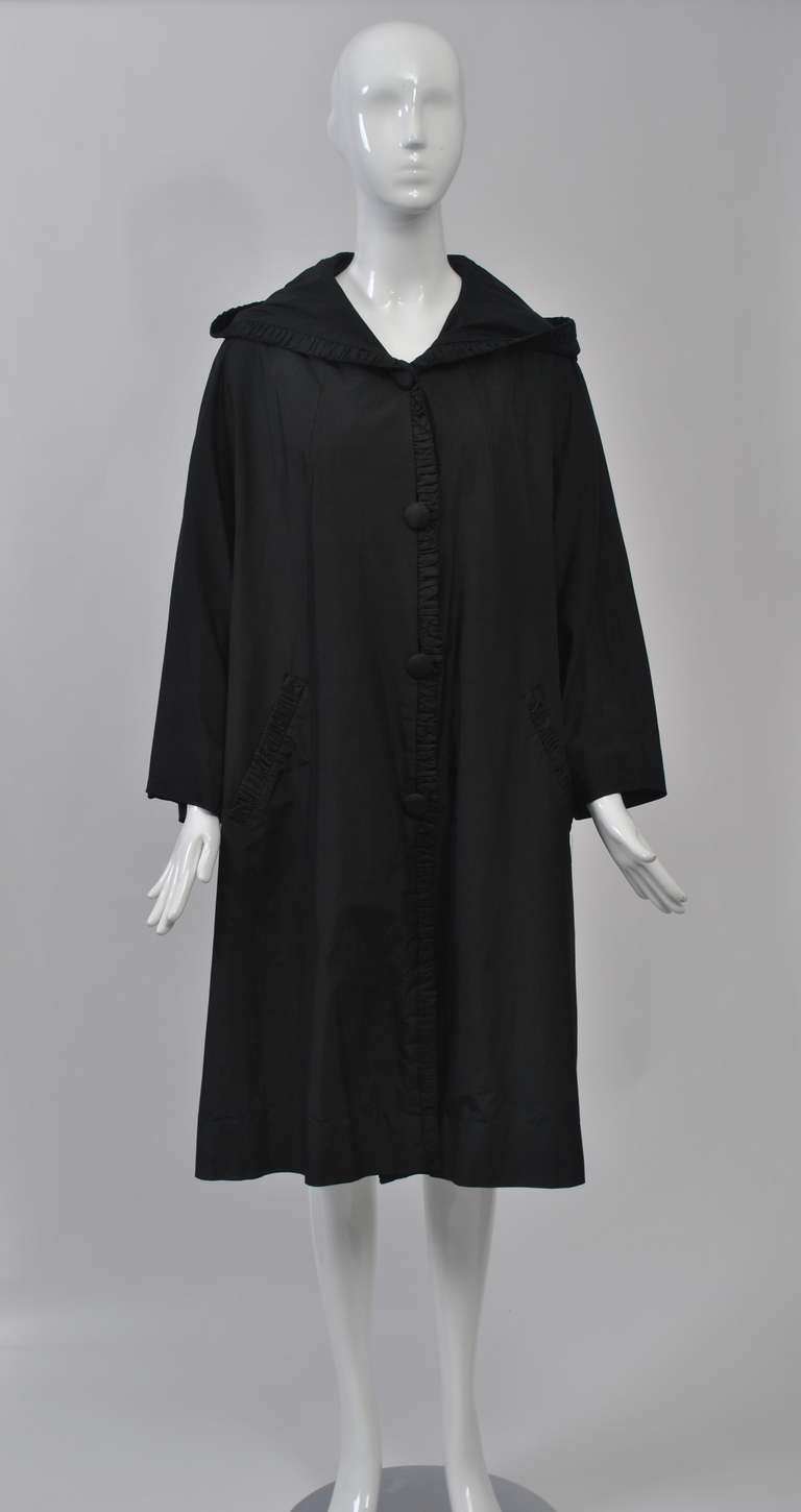 1960s black silk raincoat in swing style with pleated edging. Single breasted with wide collar/hood. Self buttons and button holes concealed in edging seam. Slash pockets with pleated edge. Raglan, wrist-length sleeves. Lightweight and versatile.