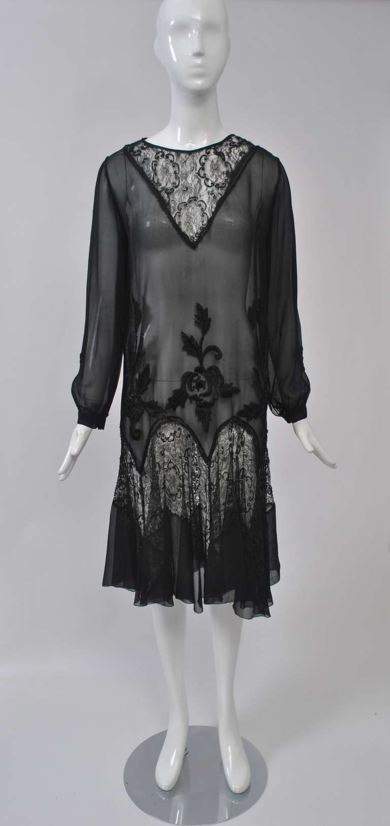 1930s chemise dinner dress of sheer black chiffon with V-shaped laced inset at neck and gored, dropped skirt combining lace and chiffon from repeating curves. Black sateen floral embroidery embellishes the hip area. Long, sheer sleeves  gathered at