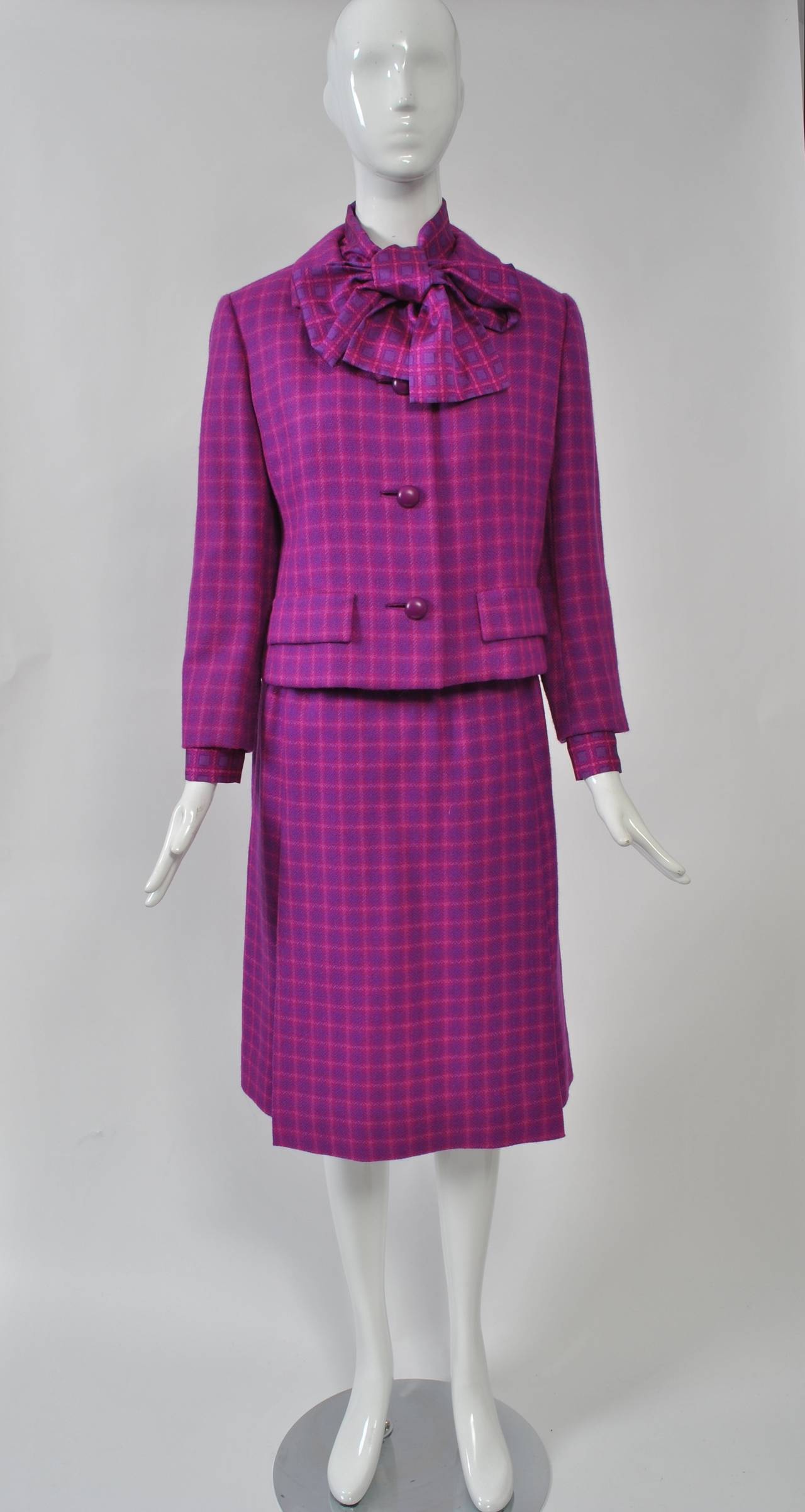 Christian Dior New York 1960s three-piece suit fashioned of fuchsia wool plaid and consisting of jacket, skirt, silk blouse, and matching hat. The single-breasted, short jacket is slightly shaped and has a notched collar and pocket flaps. Below is a