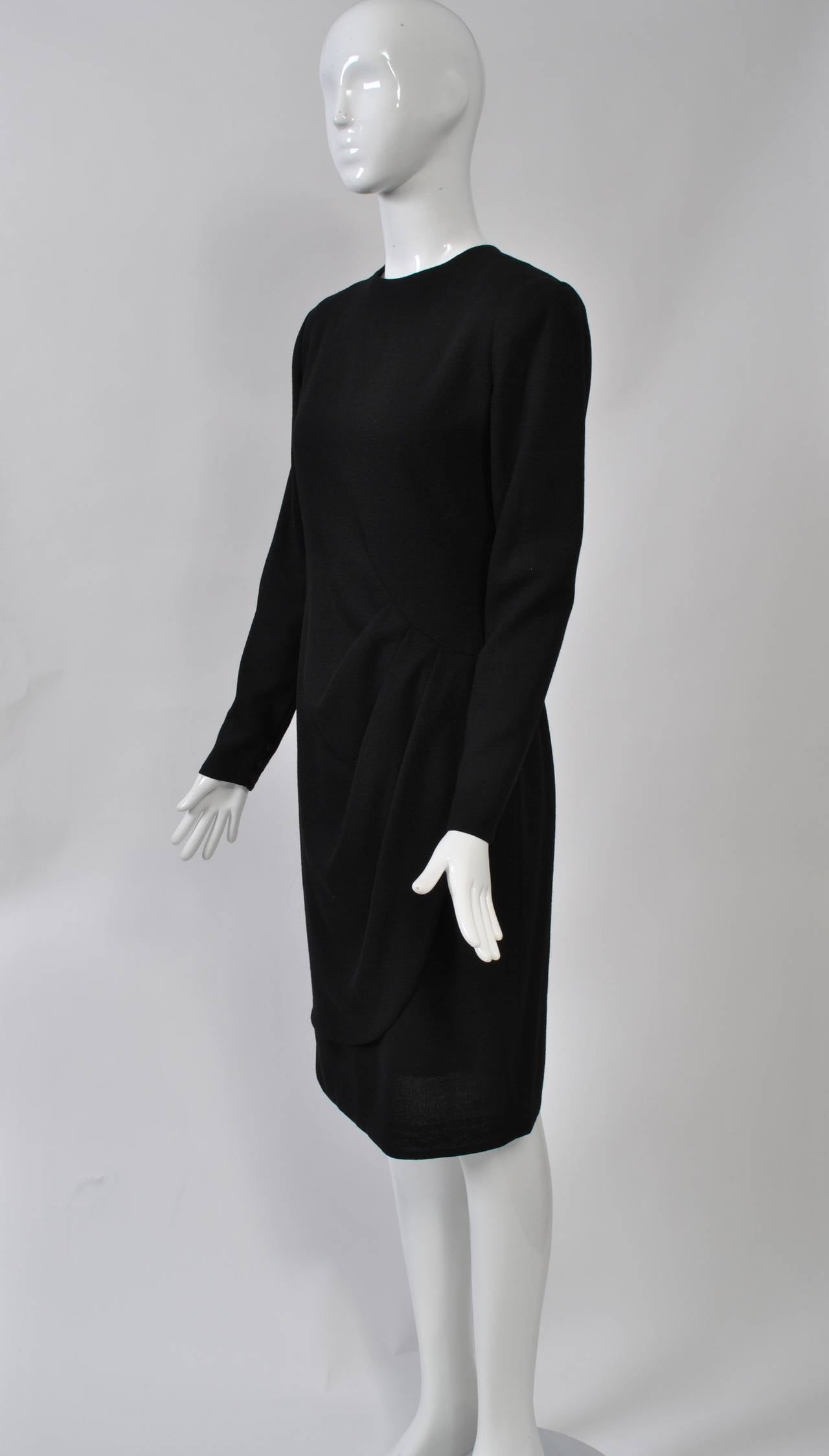 LBD by Bill Blass can take you anywhere, dressed up or down. In black wool crepe, the simple sheath dress is distinguished by a wrap front overskirt gathered on one side. High neck and long sleeves with buttoned wrists. Back zipper. Lined.