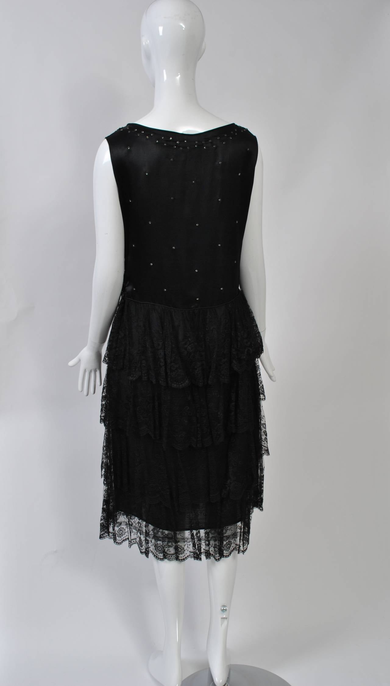 Hard-to-find 1930s cocktail dress in excellent condition with a scoop-neck, sleeveless bodice of black silk charmeuse and a tiered skirt of black lace. The bodice is studded with rhinestones and has a dropped waist that tapers to a V in front. Snaps