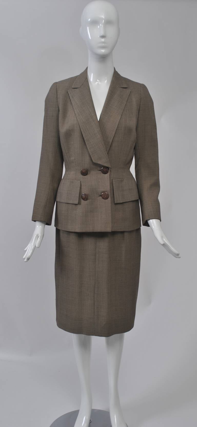 Immaculate styling is the hallmark of this 1950s suit fashioned of a fine brown and white tweed wool. The long blazer-style jacket features a notched shawl collar cut diagonally at bottom, large pocket flaps at the hips and double-breasted buttons