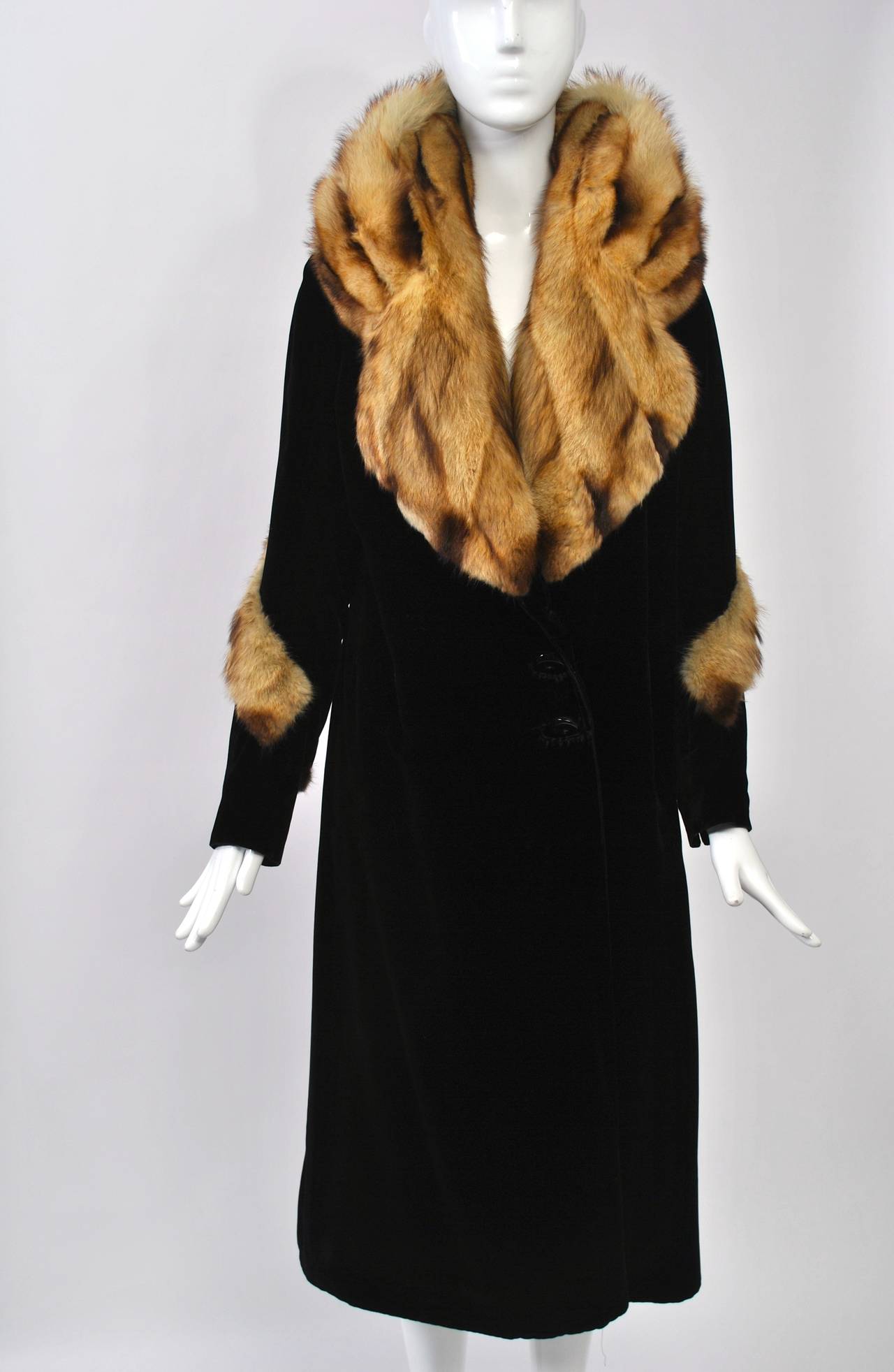 Art Deco evening coat of black velvet featuring a huge fur collar, possibly fitch, that curves up into the neck before settling on the shoulders. Narrow, straight cut to the coat and sleeves, which are trimmed with strips of the fur diagonally