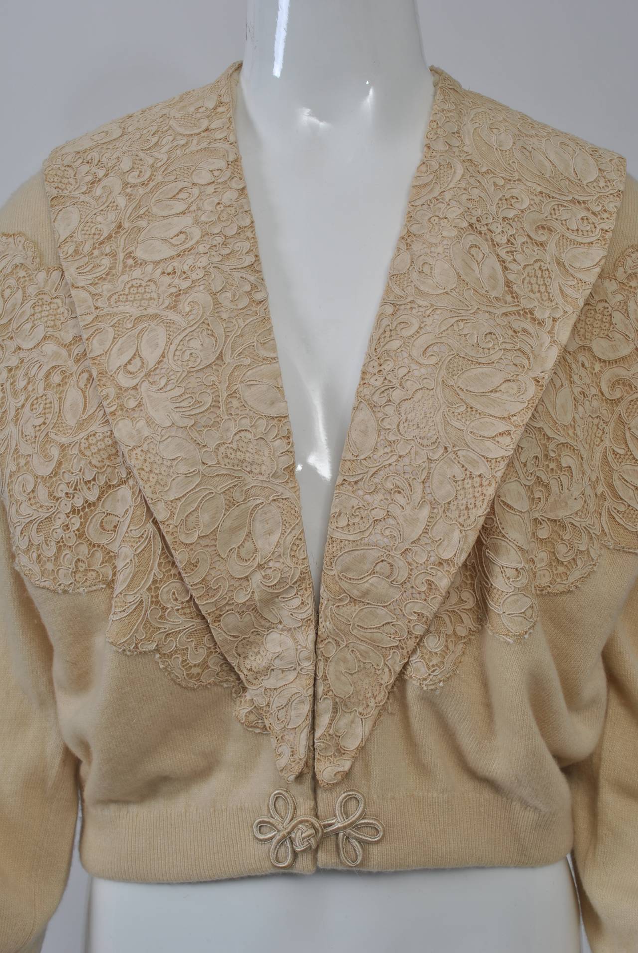 An unusual version of the decorated sweaters from the 1950s-'60s, many of which have a detachable fur collar, this beige cashmere cardigan has lace trim and a lace shawl collar that snaps on and off. Frog closure at waist and satin buttons at