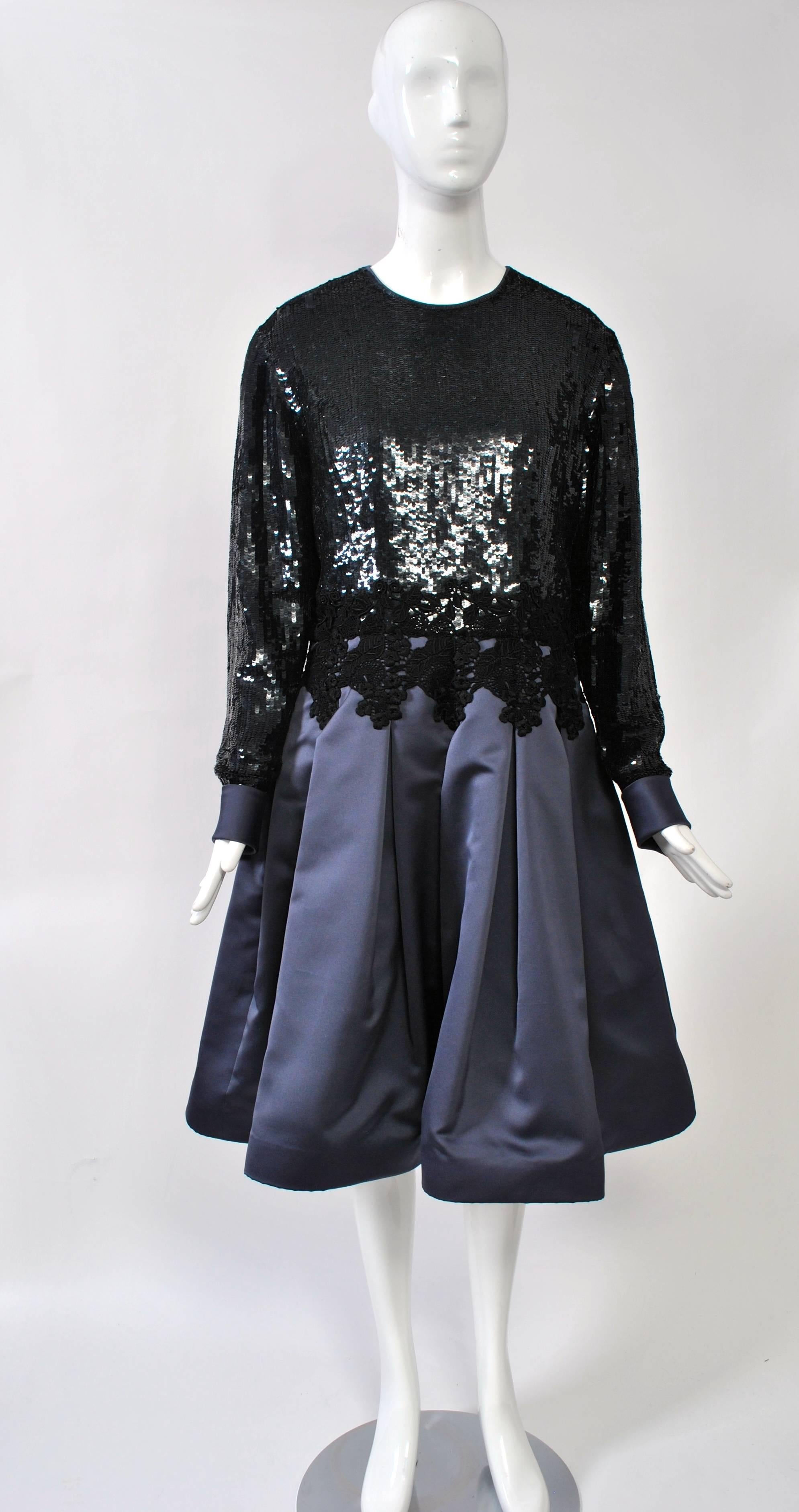 Cocktail dress featuring an all-over black sequin bodice and a gunmetal satin skirt, the bodice with jewel neckline and long sleeves, the satin skirt with box pleats sewn down with a repeating embroidery appliqué below the waist. Satin cuffs with