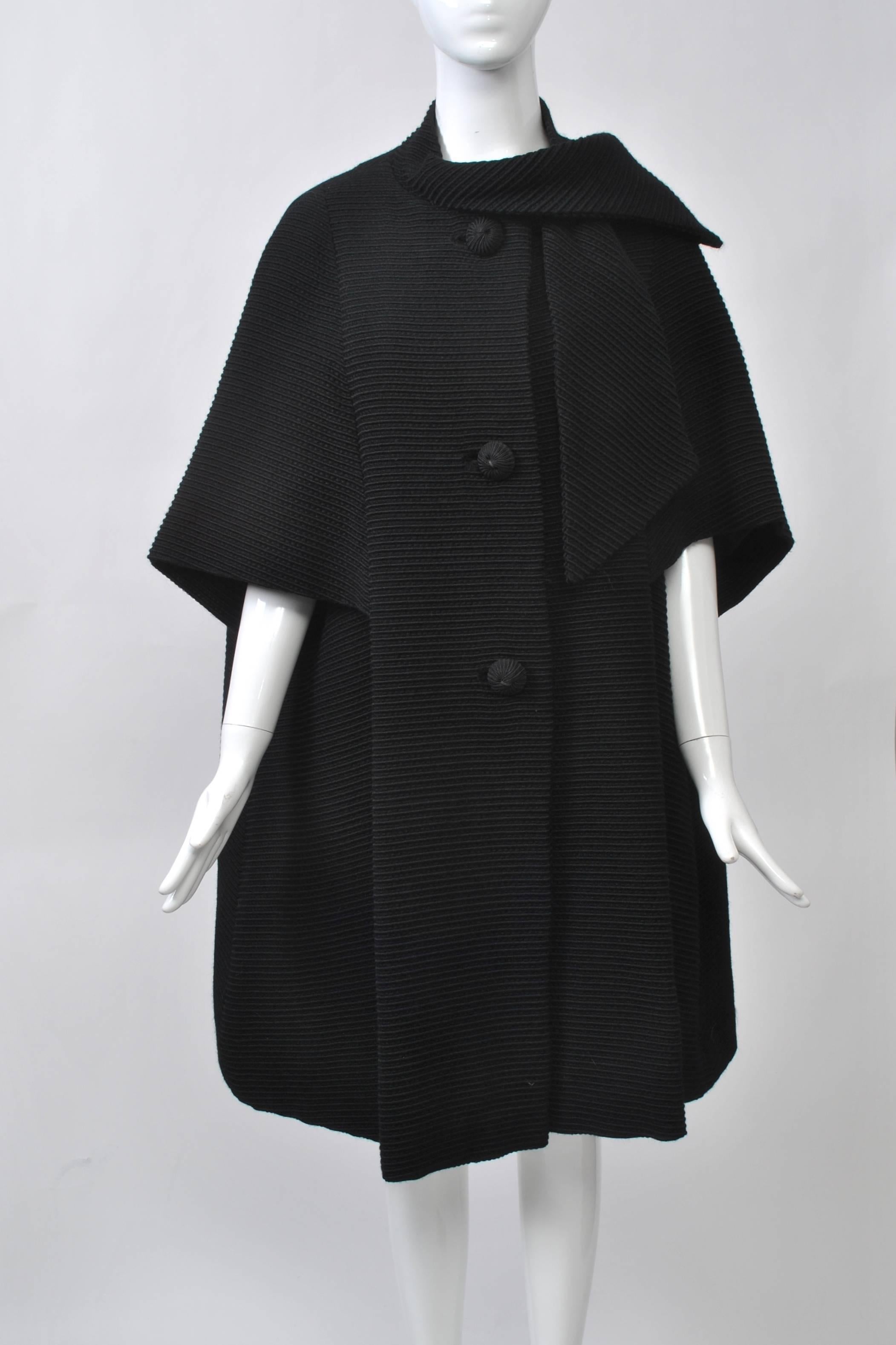 Stunning 1960s coat with elbow-length cape sleeves in heavy black ottoman-ribbed wool. Swing style with three large cord buttons and self tie at neck. Black satin lining is interlined for warmth. Unlabeled. Fits S-M.