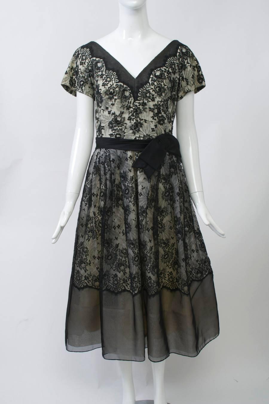Black lace and organza dress over a white lining features a fitted bodice and full skirt, short sleeves, V neck front and back, and a black waist sash with bow. The edges of the lace are scalloped and trimmed with sheer organza at the hem and