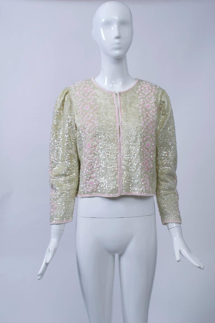 1980s hook-front cardigan allover sequined in iridescent off-white accented by pink beading around the borders and in a vertical floral pattern front and back and down the sleeves. Shirred shoulders, shoulder pads, lined.
