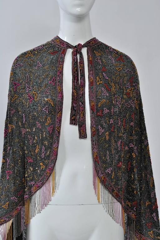 Exquisite and rare 1930s cape featuring multicolor microbeading on a black chiffon ground in an allover squiggle design. Finished around hem with matching beaded fringe. Narrow neckband continues into tie front. Drapes beautifully. Accompanied by