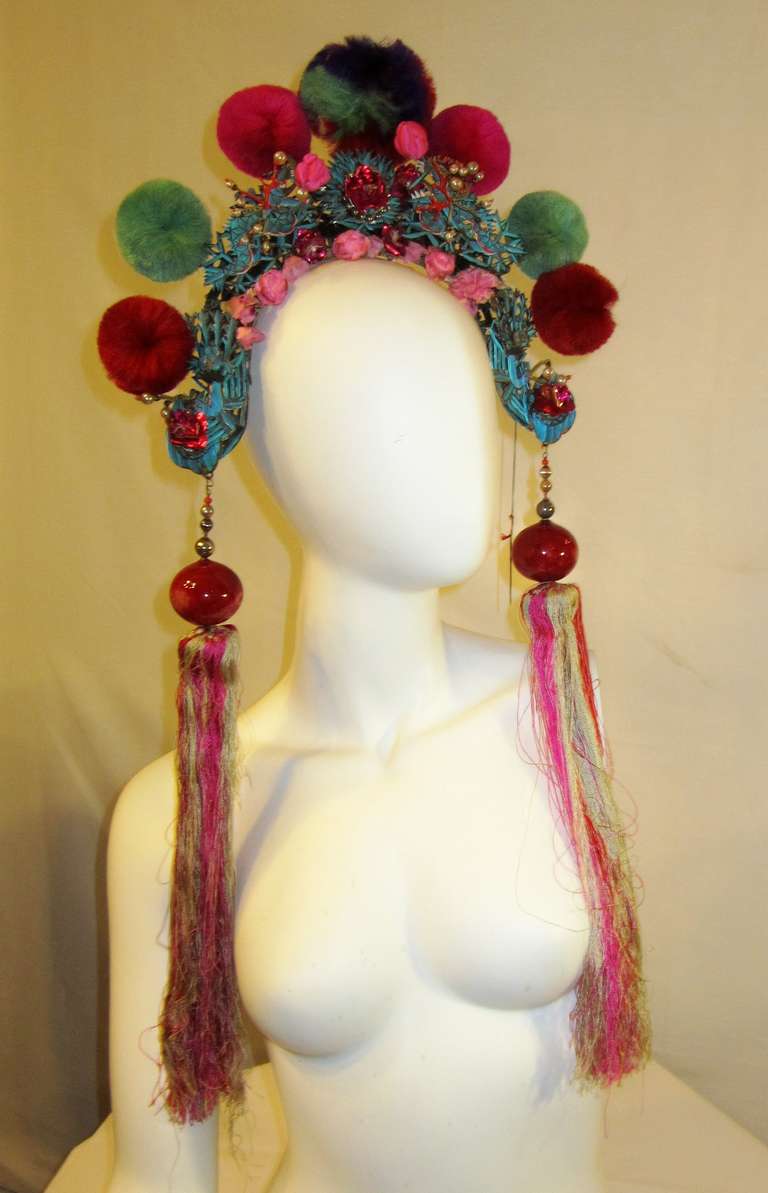 This is a wonderful 20th century Chinese wedding/theater headdress comprised of pom-poms in cranberry red, turquoise, and purple on a thick board and decorated with pink silk flowers, violet tin foil flowers, faux pearls and faux branch coral