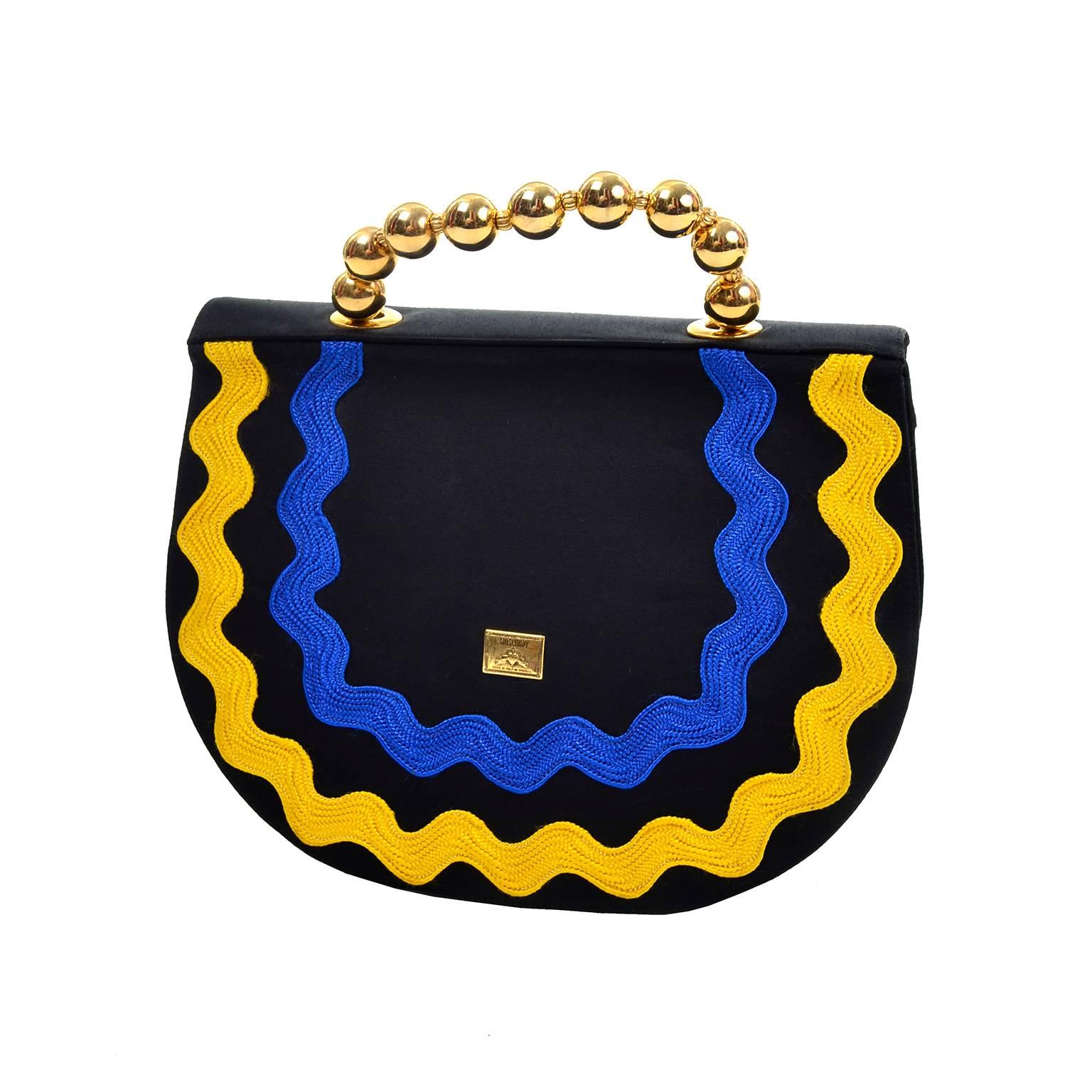 This is a really fun black fabric handbag by Moschino with wavy arching lines of rick rack following the curve of the bag in green, magenta, yellow and blue. These rope-like ribbons are thick, creating a relief on the bag. The handle is made of