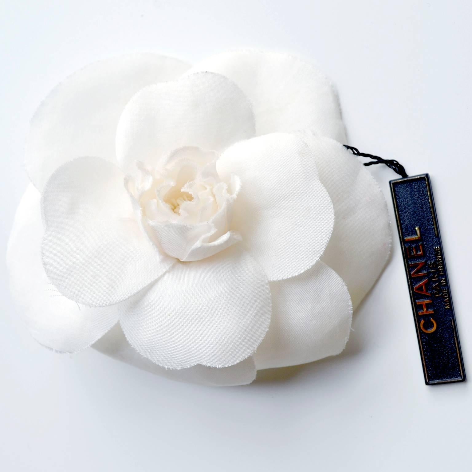 This original Chanel Camelia silk brooch or lapel pin comes with its tags still attached and in its original box.  This lovely brooch is ivory and could be worn in a number of ways!  The Chanel gold tone plate reads: 